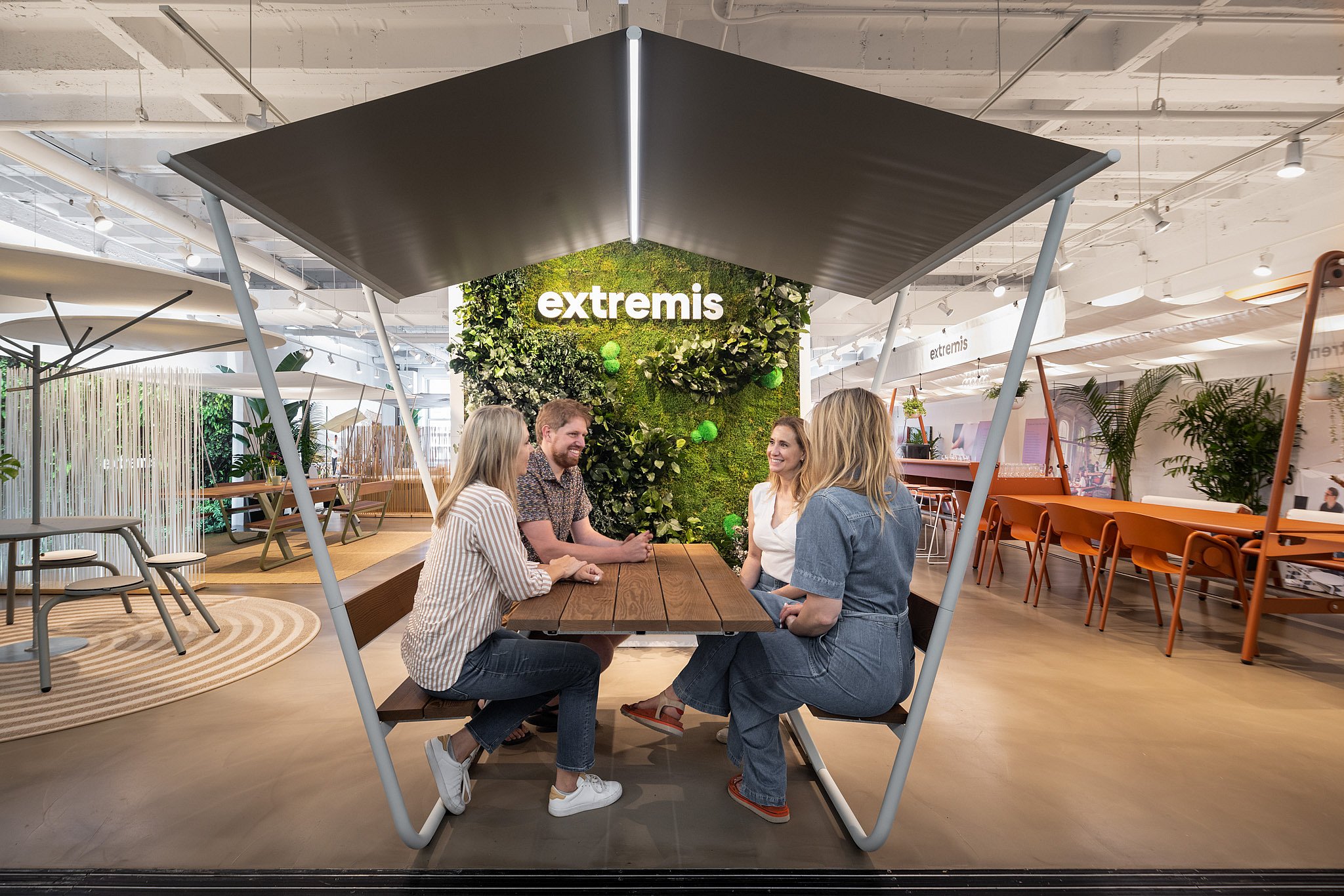 Vier Personen sitzen im NeoCon-Showroom an einem hölzernen Manila-Tisch unter einem großen, eckigen Vordach mit einer grünen Pflanzenwand und einem leuchtenden Extremis-Logo dahinter.