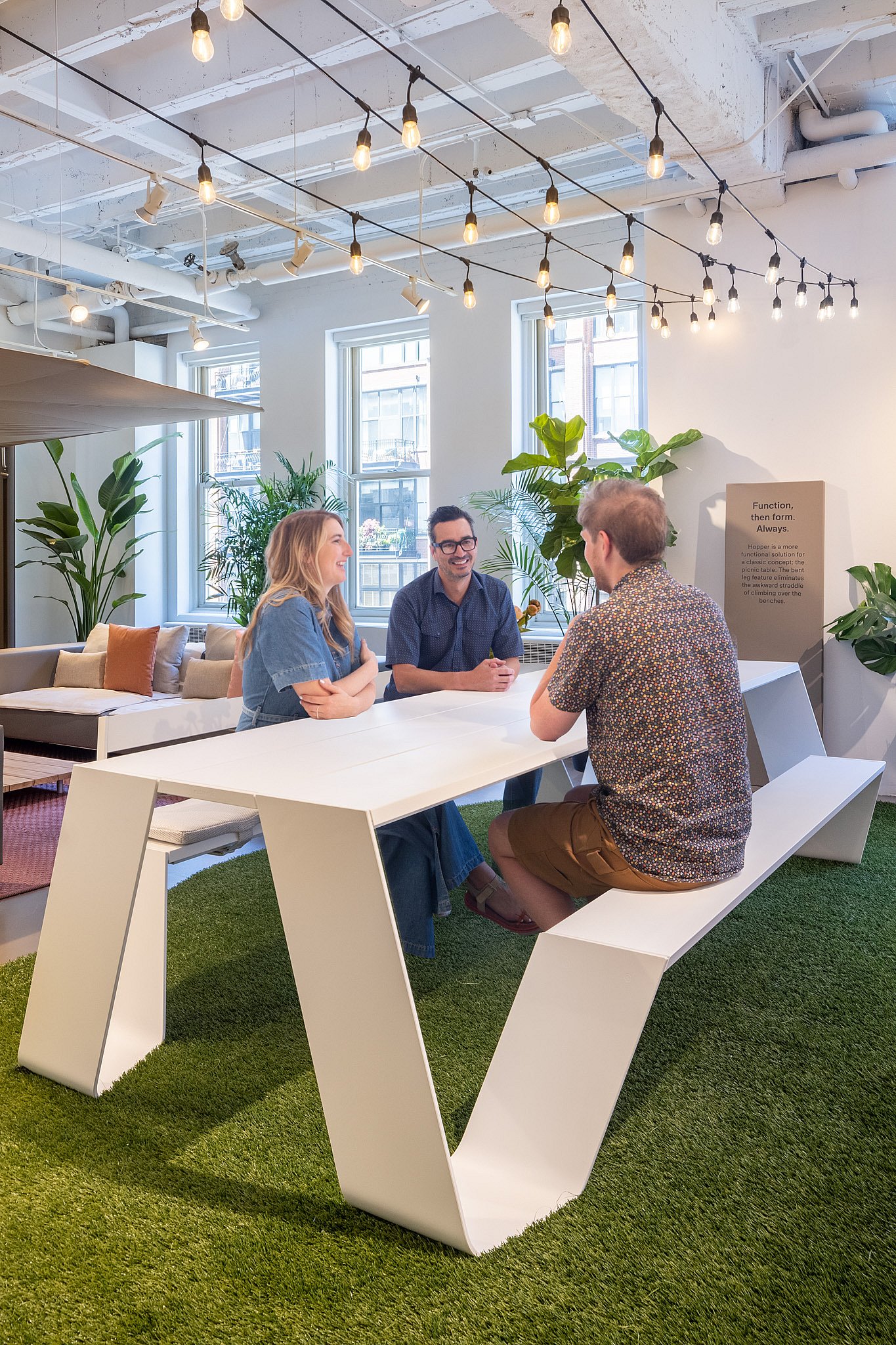 Drei Personen unterhalten sich um einen großen weißen Trichtertisch auf Kunstrasen unter Lichterketten, umgeben von Pflanzen und Loungemöbeln in einem hellen Indoor-Showroom.