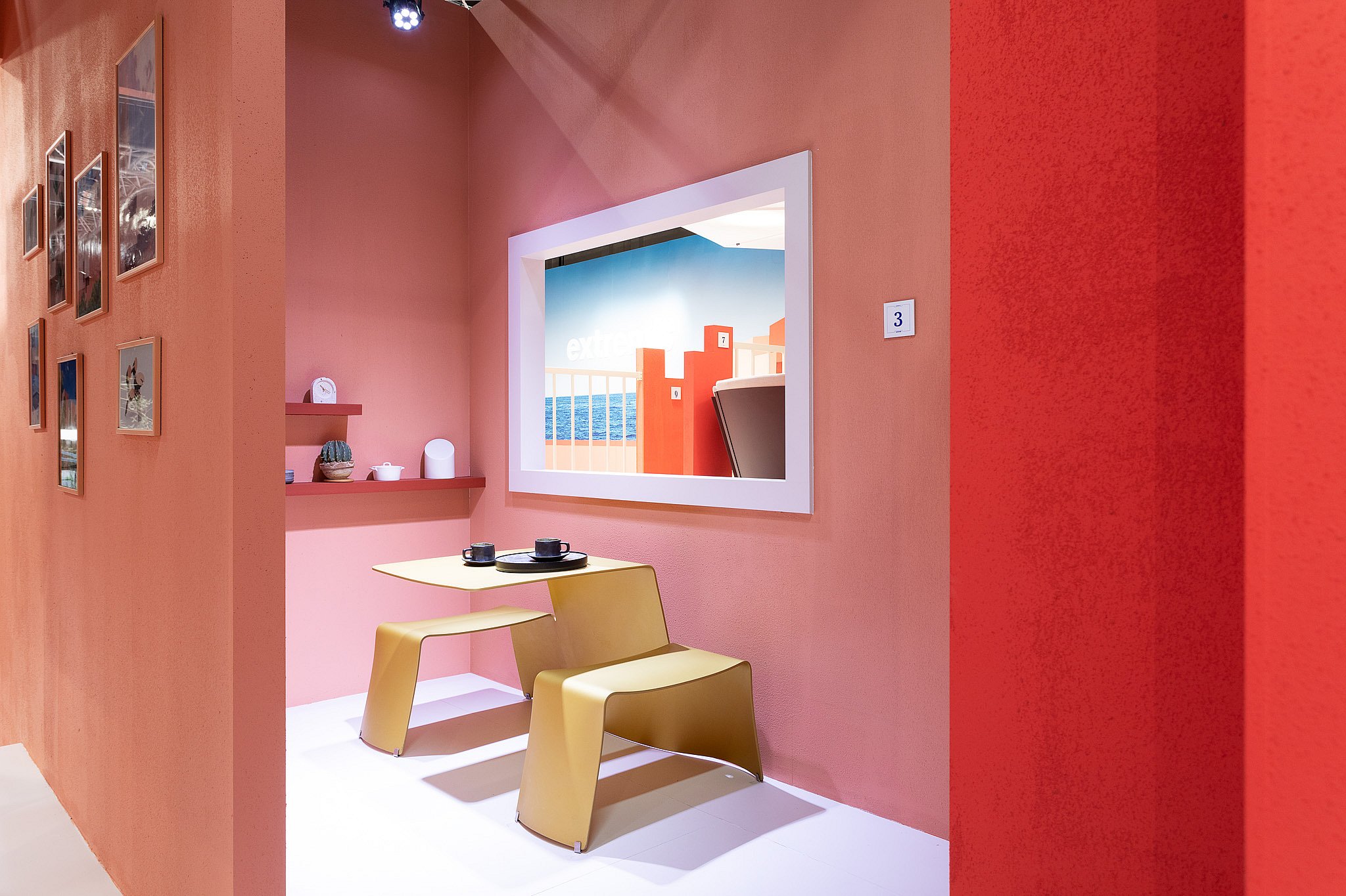 Small café-like corner with coral walls, a yellow table and benches, decorative shelves, and a framed opening looking out toward the exhibition stand.