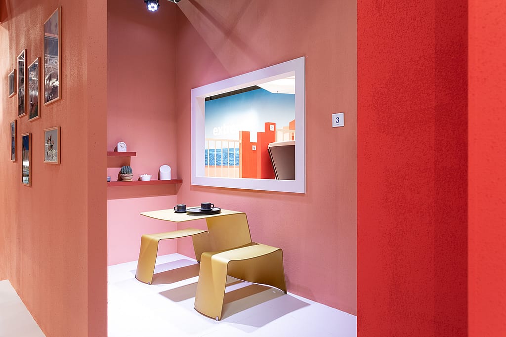 Small café-like corner with coral walls, a yellow table and benches, decorative shelves, and a framed opening looking out toward the exhibition stand.