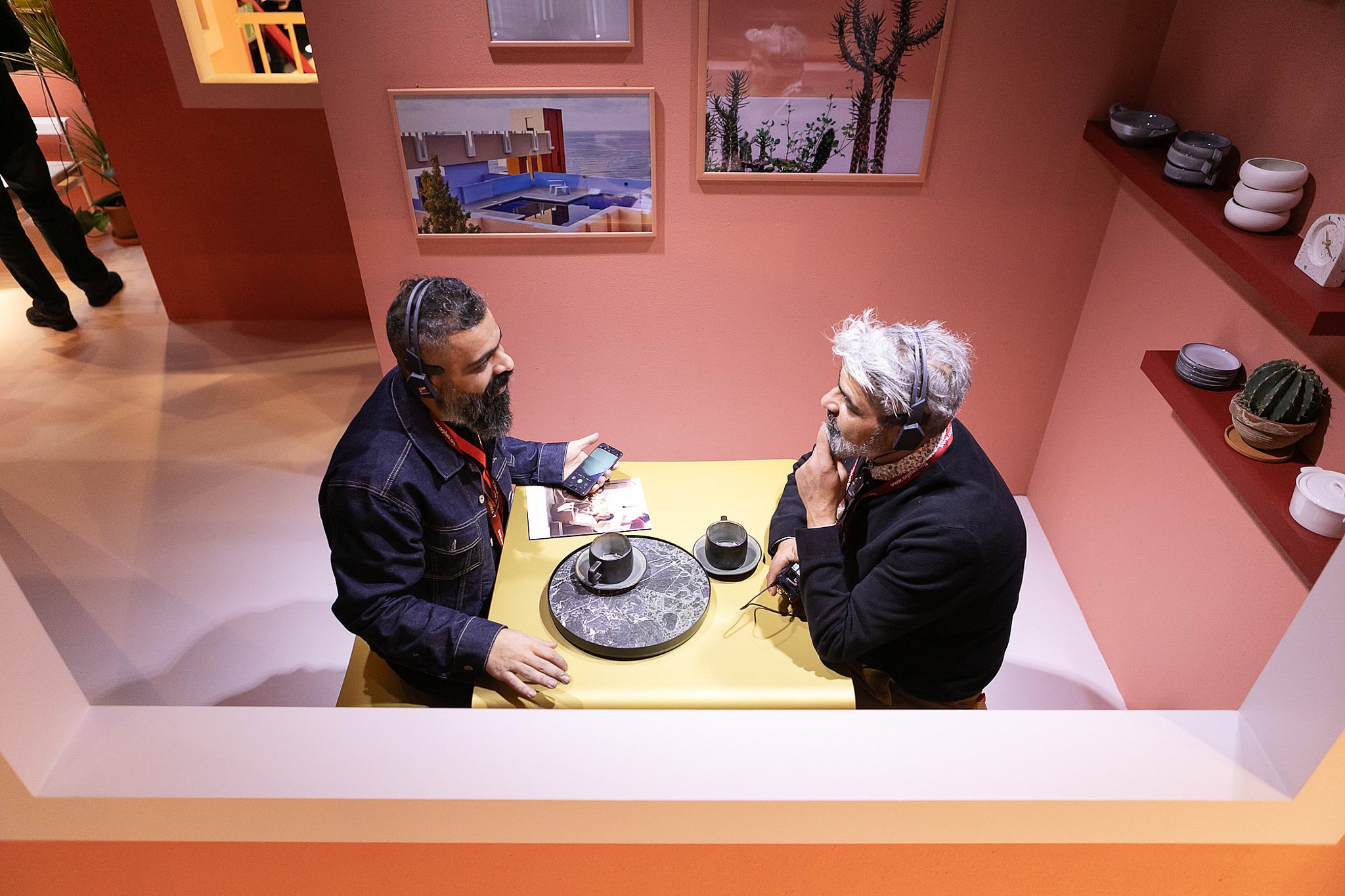 Overhead view of two visitors wearing headphones as they sit across from each other at a small yellow picnik table with coffee cups and a round tray.