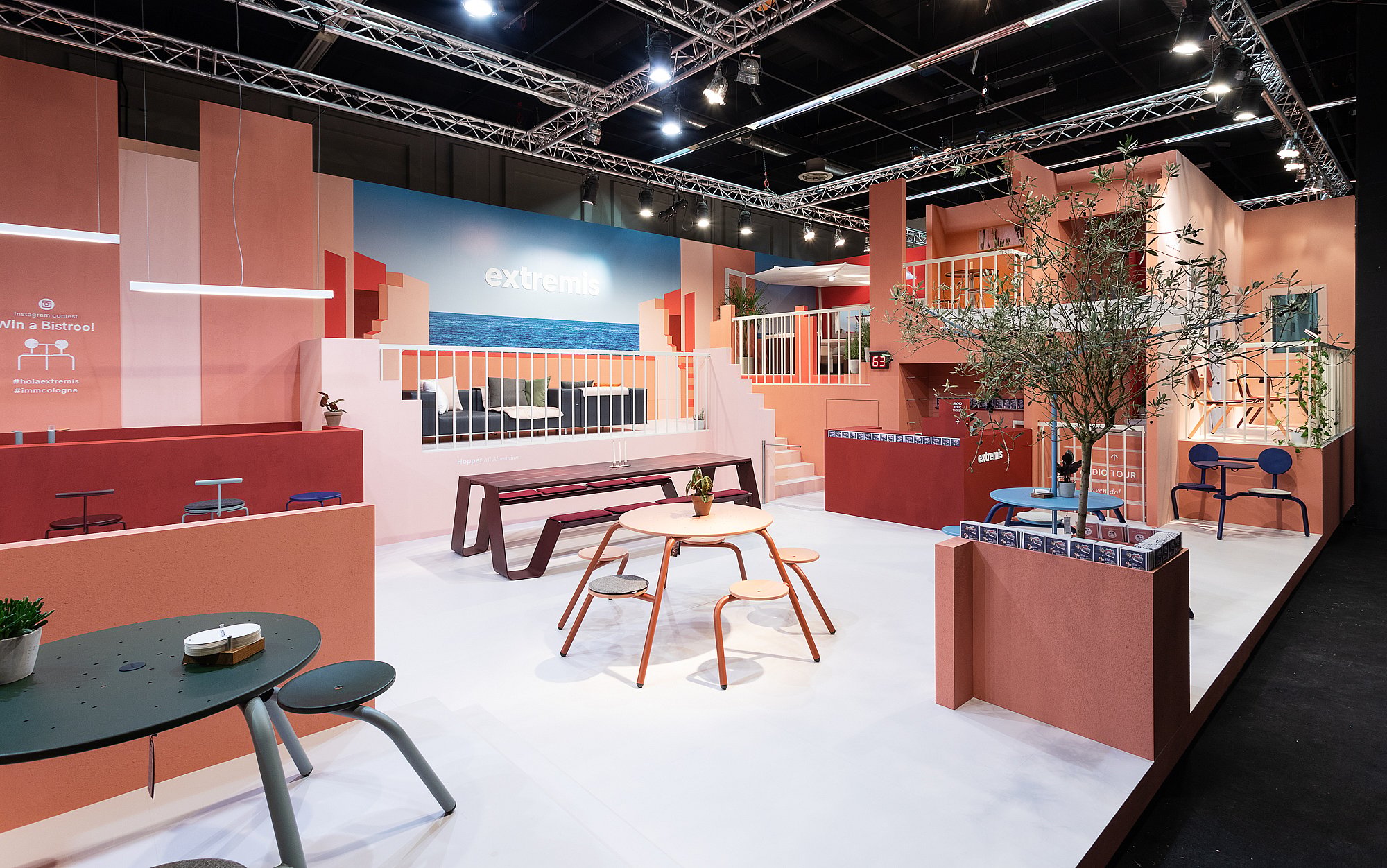 Two people sit facing each other at a compact yellow picnik table with matching benches inside a pink exhibition nook, drinking coffee beneath a large interior window.