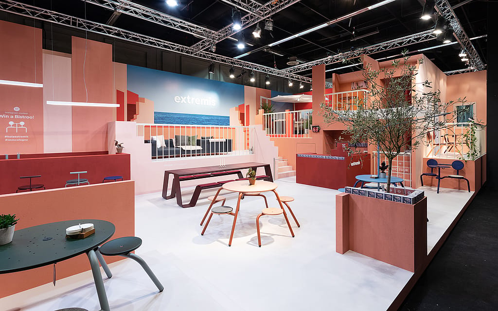 Two people sit facing each other at a compact yellow picnik table with matching benches inside a pink exhibition nook, drinking coffee beneath a large interior window.