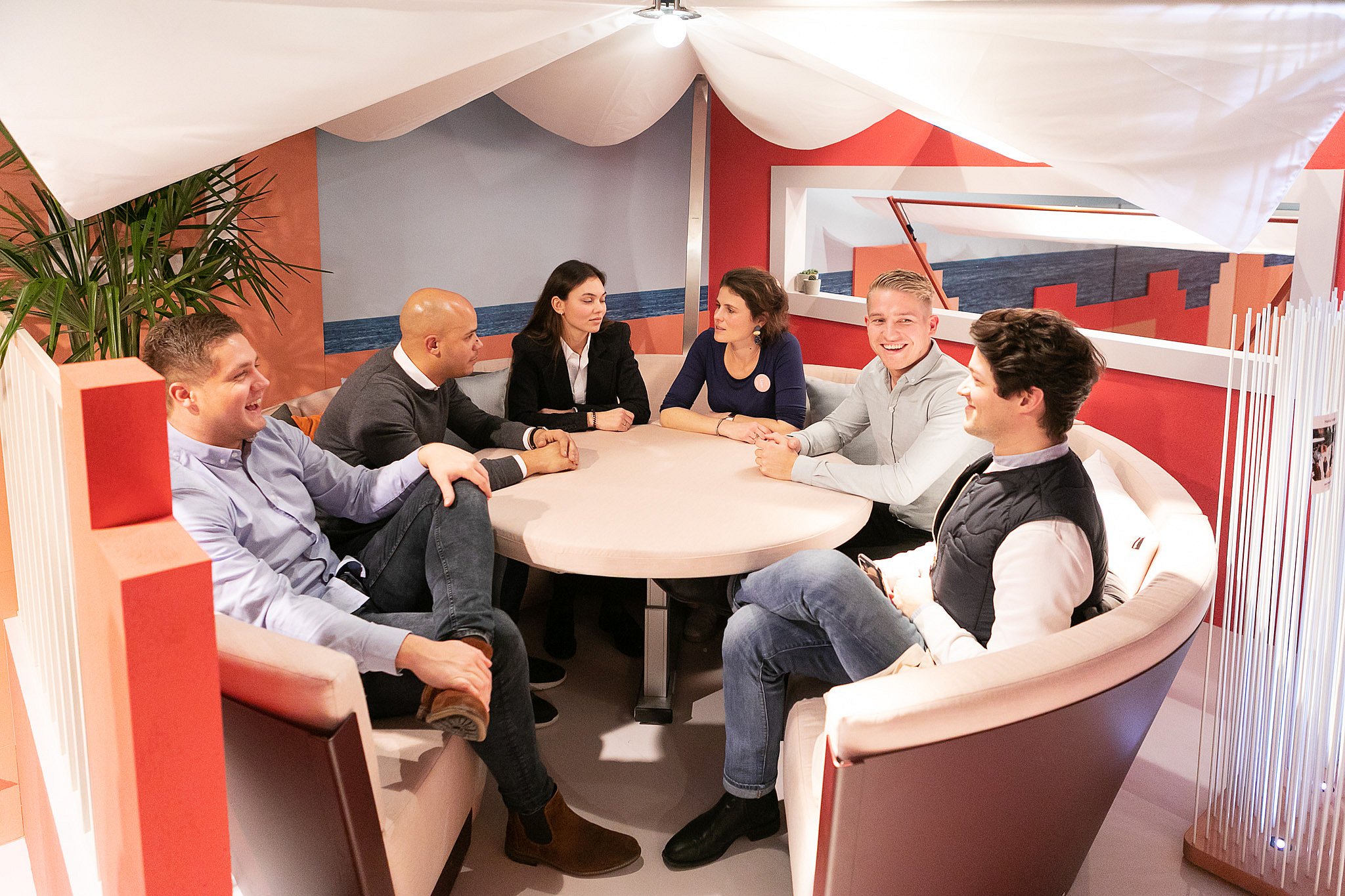 Six people sit together around a round table inside a circular Kosmos seating unit beneath draped white fabric, talking in a lounge-like setting.