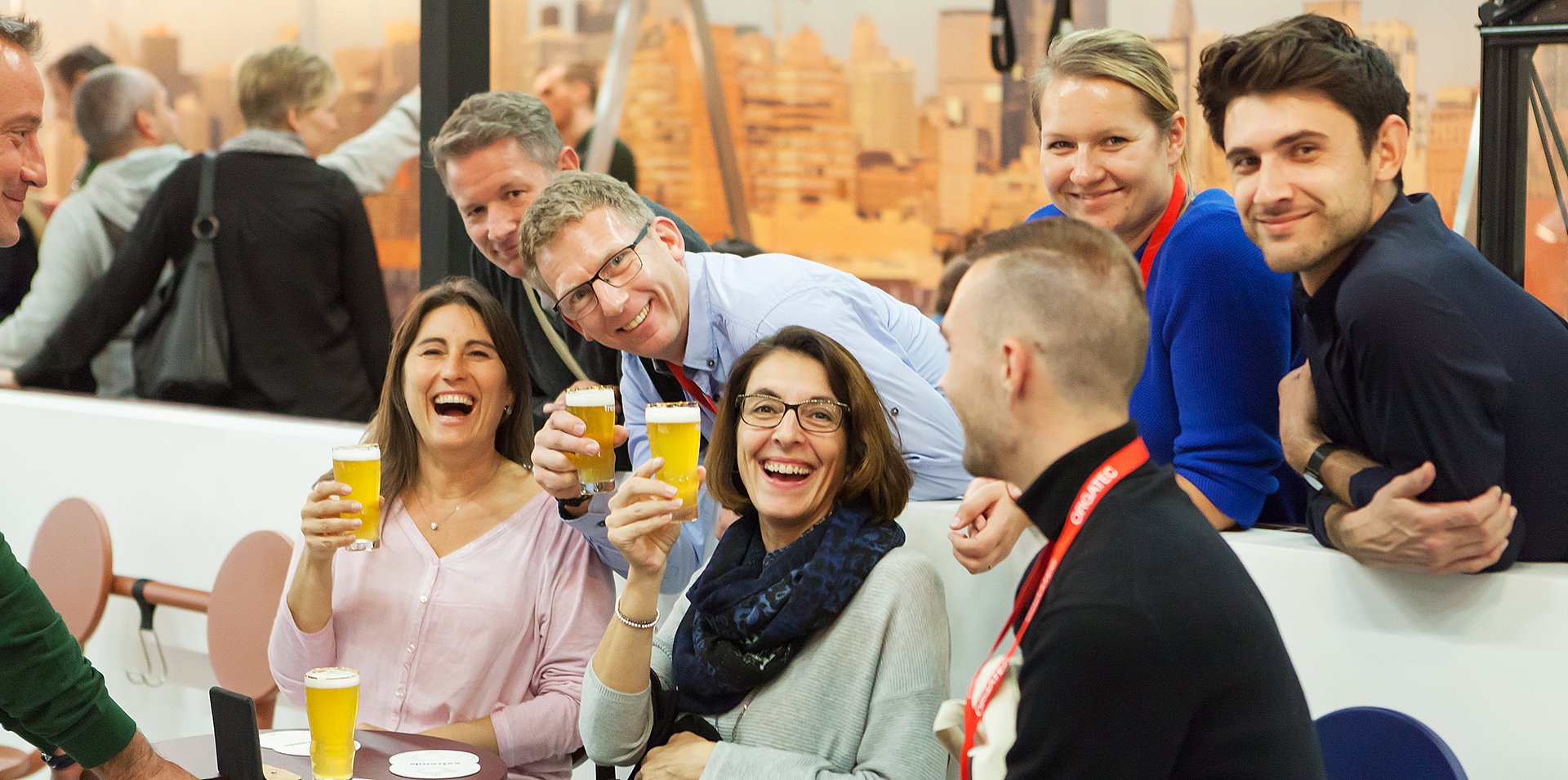 Un groupe de visiteurs souriants se penchent ensemble et lèvent des verres de bière au-dessus d’une table compacte, créant ainsi un moment social animé sur le stand de la foire.
