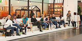 Les visiteurs s’assoient et discutent le long d’un long banc blanc équipé de tables compactes Bistroo de différentes couleurs, avec des ampoules suspendues et des structures de serre derrière elles.