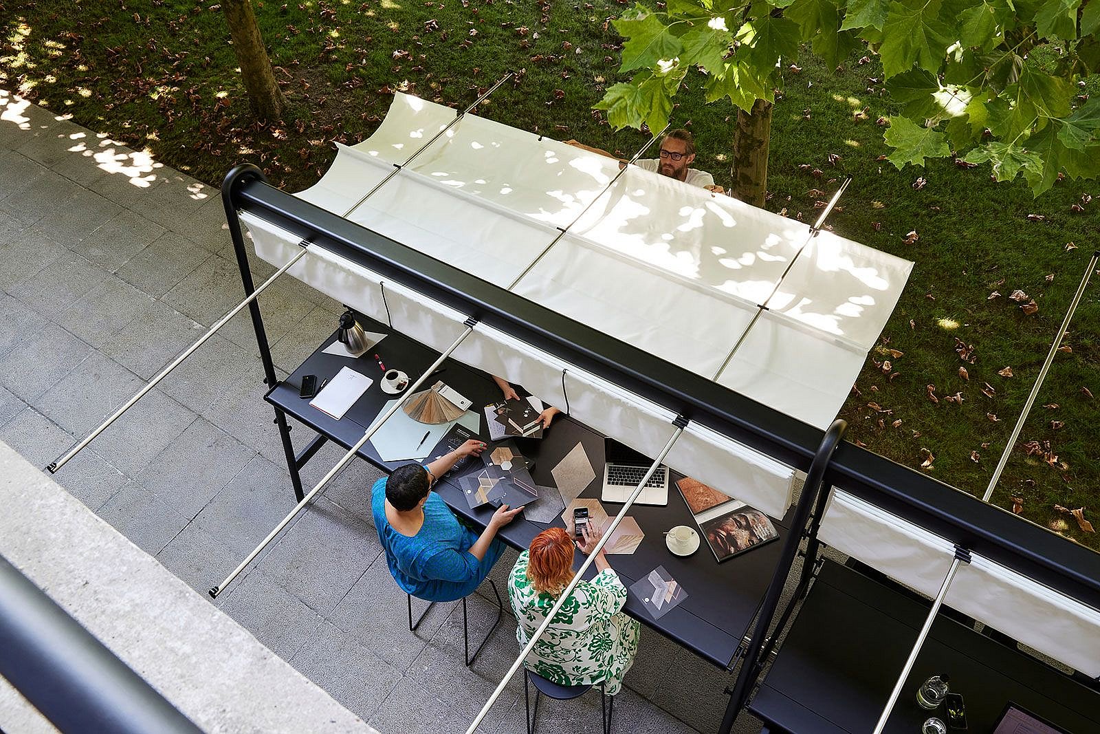 Overhead view of two coworkers reviewing material samples at a long outdoor table under an extended white canopy, while another person adjusts the shade from the lawn side.