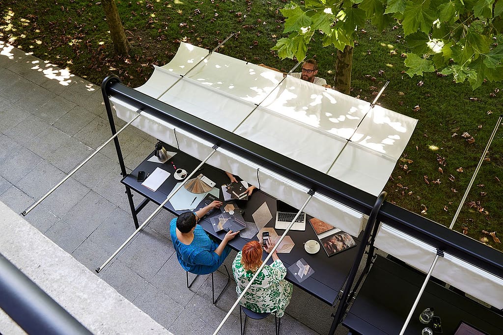 Overhead view of two coworkers reviewing material samples at a long outdoor table under an extended white canopy, while another person adjusts the shade from the lawn side.