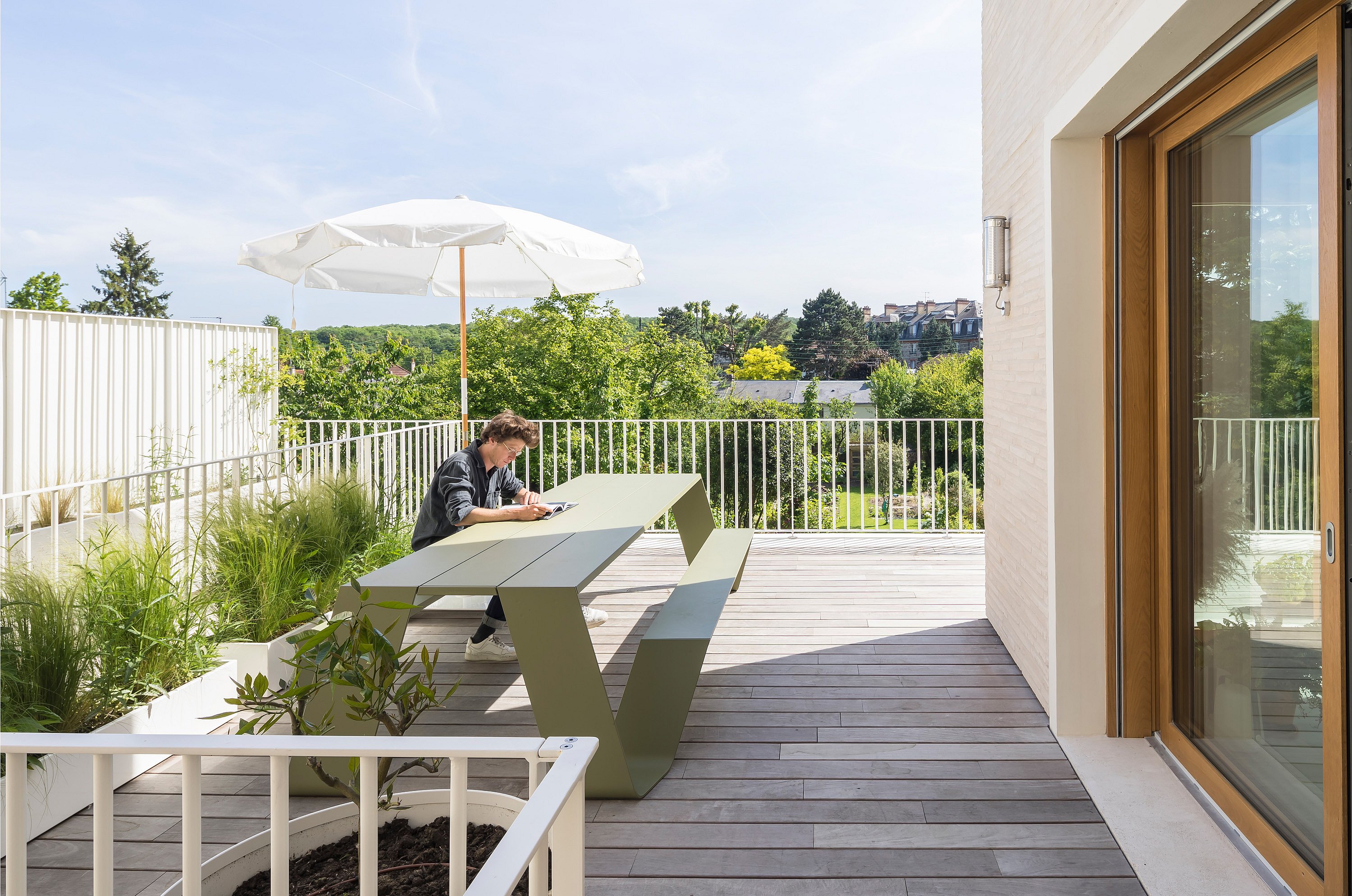 Die Person arbeitet allein an einem langen grünen Picknicktisch im Freien auf einer Dachterrasse, der von einem weißen Sonnenschirm beschattet und von bepflanzten Behältern umgeben ist.