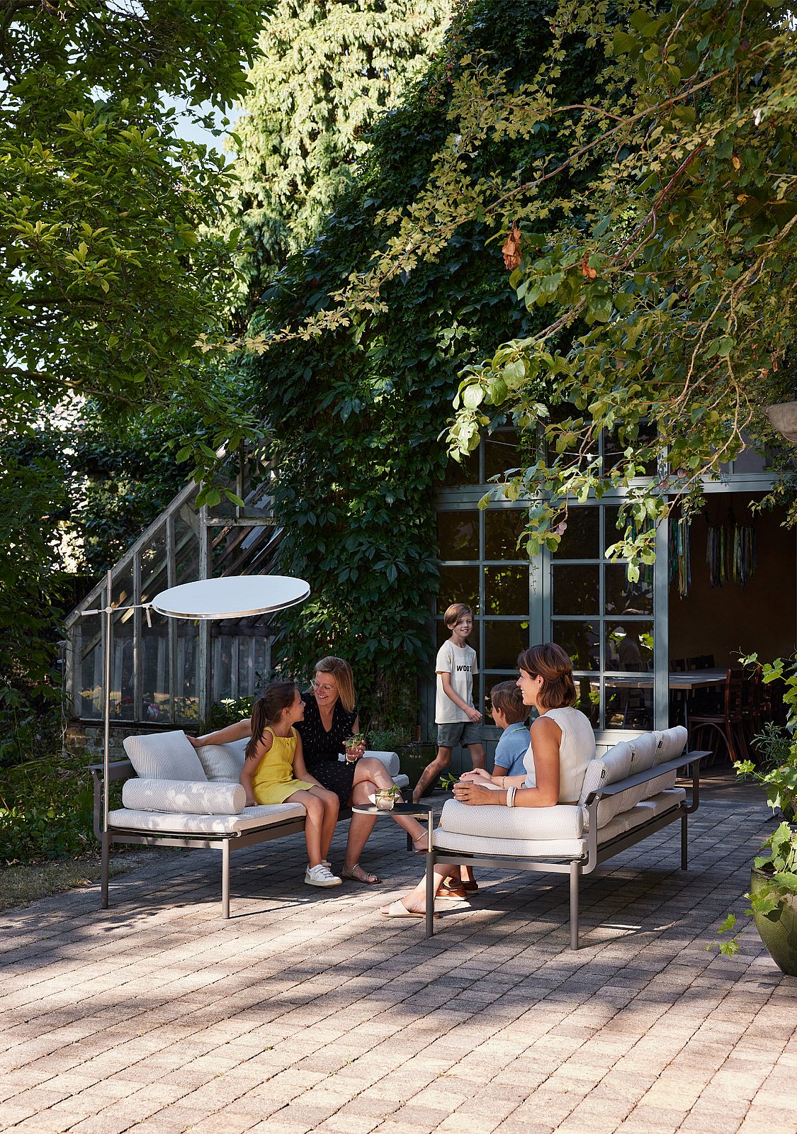   Eine Familie entspannt sich in einer hellen Lounge im Freien in einem grünen Innenhof neben einem Gewächshaus. Erwachsene und Kinder sitzen in der Nähe.