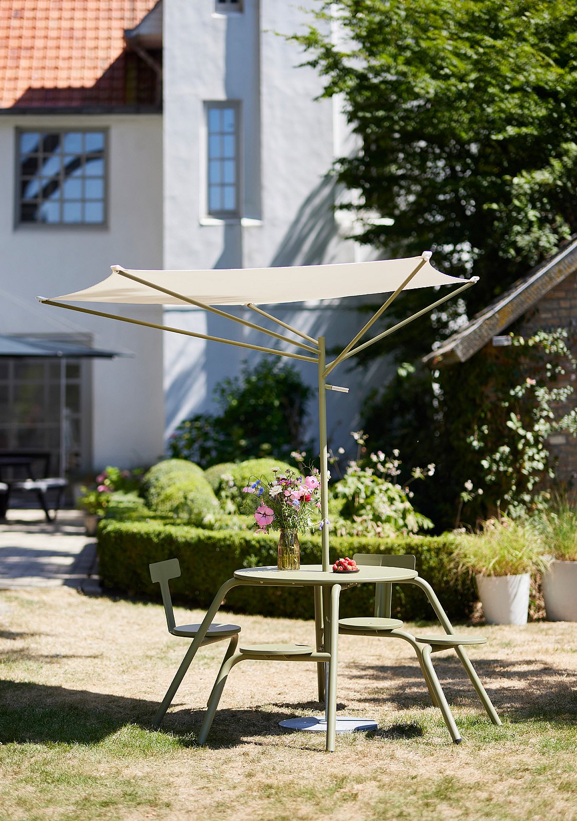 Kleiner grüner Outdoor-Tisch mit zwei passenden Stühlen steht in einem Garten unter einem cremefarbenen eckigen Sonnenschirm, mit Blumen in einer Vase auf der Tischplatte.