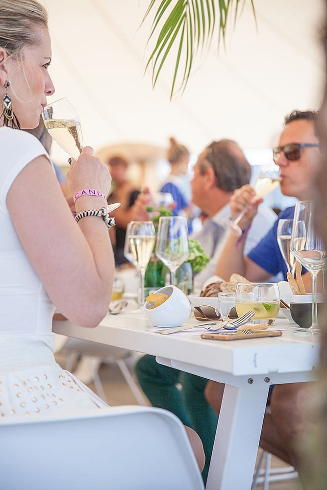   Des festivaliers sont attablés à une table blanche, dégustant du vin pétillant. Du pain, des verres et des assiettes sont disposés sur la table.