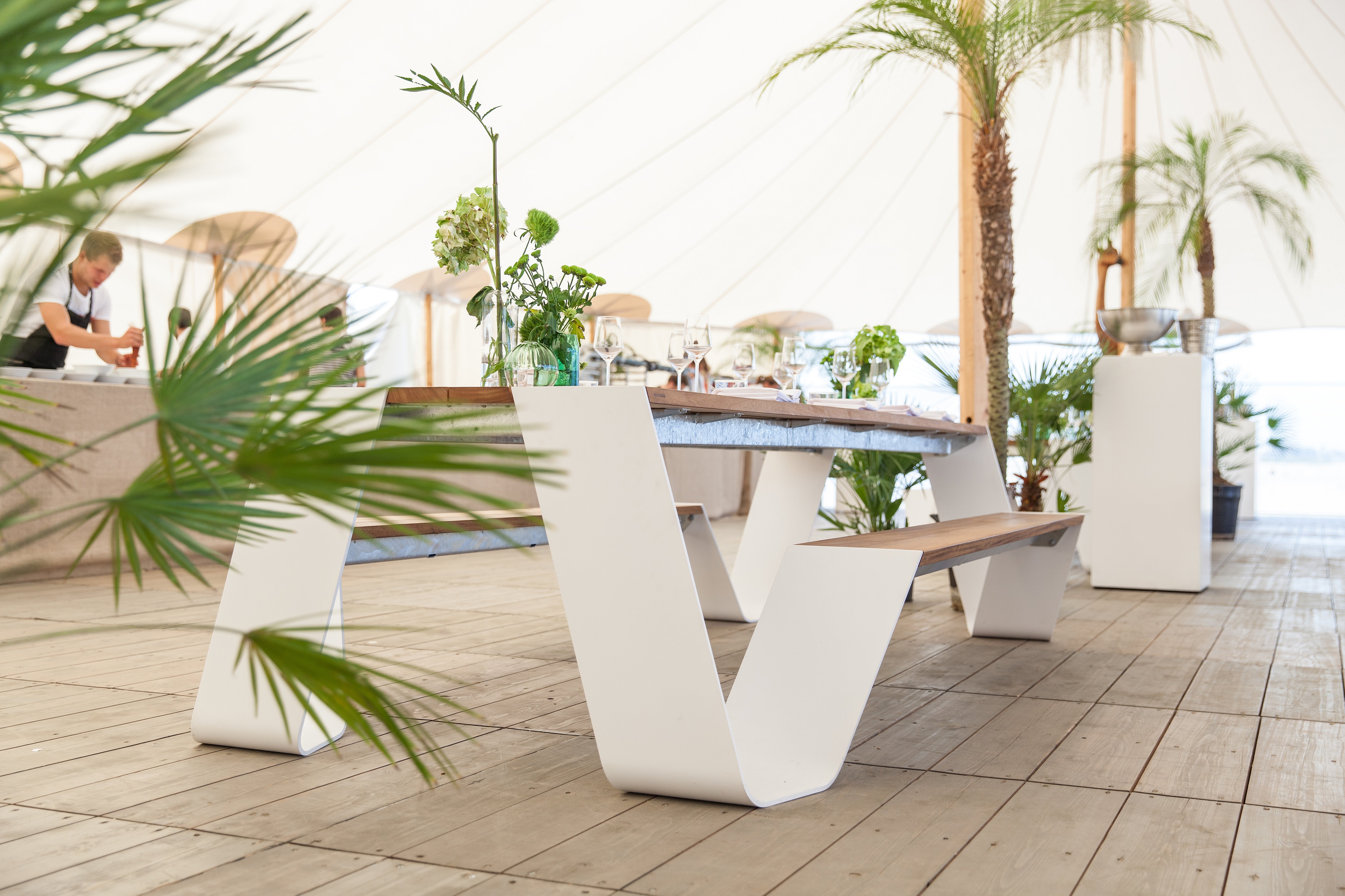 Witte buitentafel en bank op een houten vloer in een lichte festivaltent, gedecoreerd met bloemen, glaswerk en tropische planten.