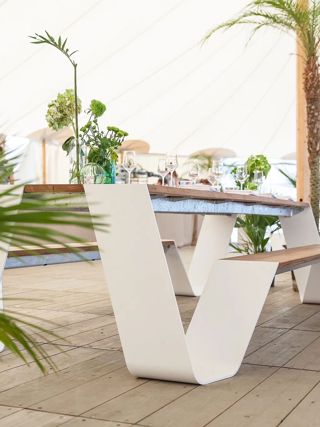 Witte buitentafel en bank op een houten vloer in een lichte festivaltent, gedecoreerd met bloemen, glaswerk en tropische planten.