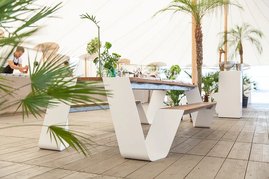 White outdoor table and bench set on a wooden floor inside a bright festival tent, styled with flowers, glassware and tropical plants.