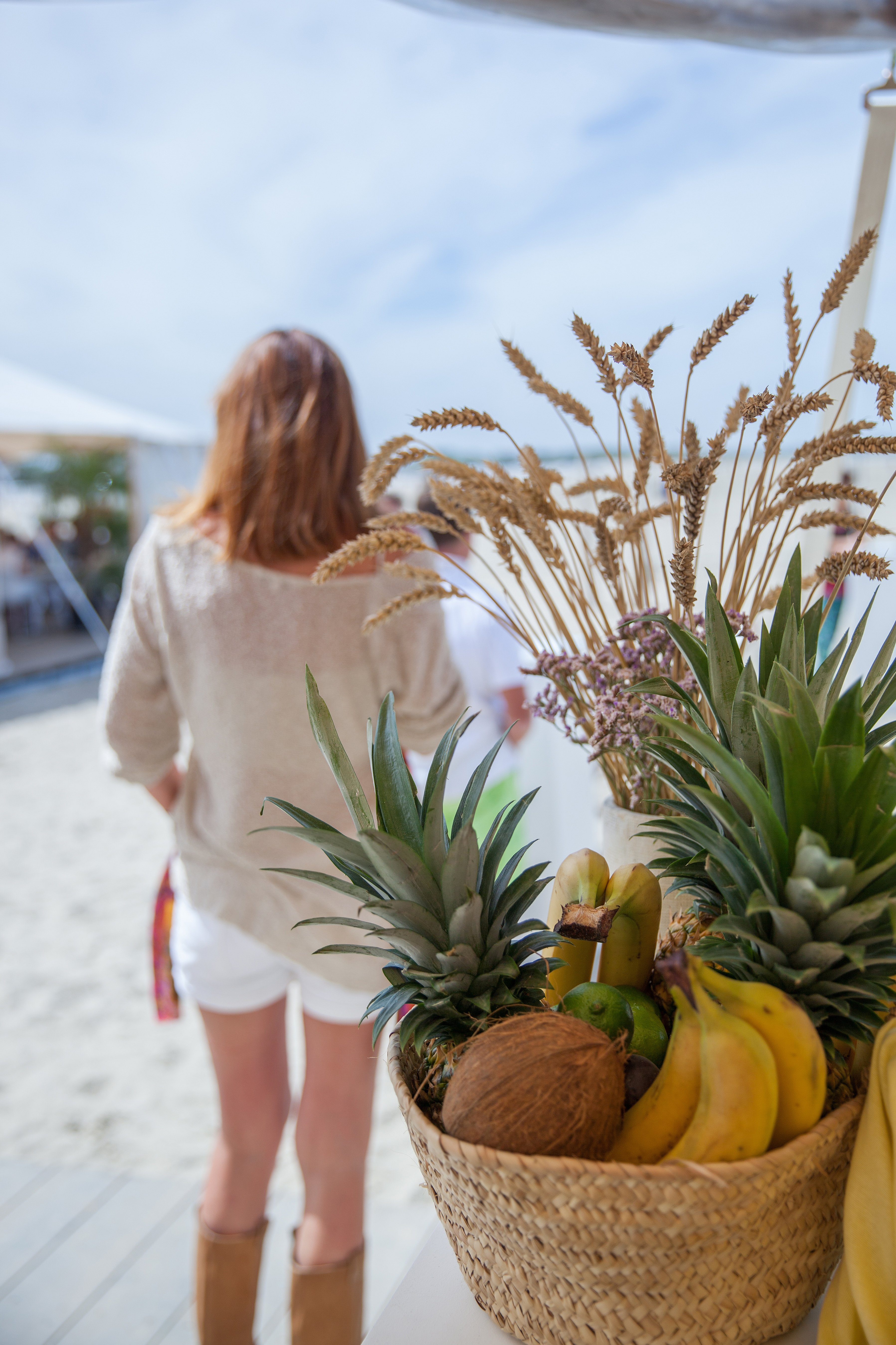   Un panier rempli d'ananas, de bananes, de noix de coco et de fleurs est exposé au premier plan, tandis que des festivaliers se dirigent vers la tente de plage située derrière.