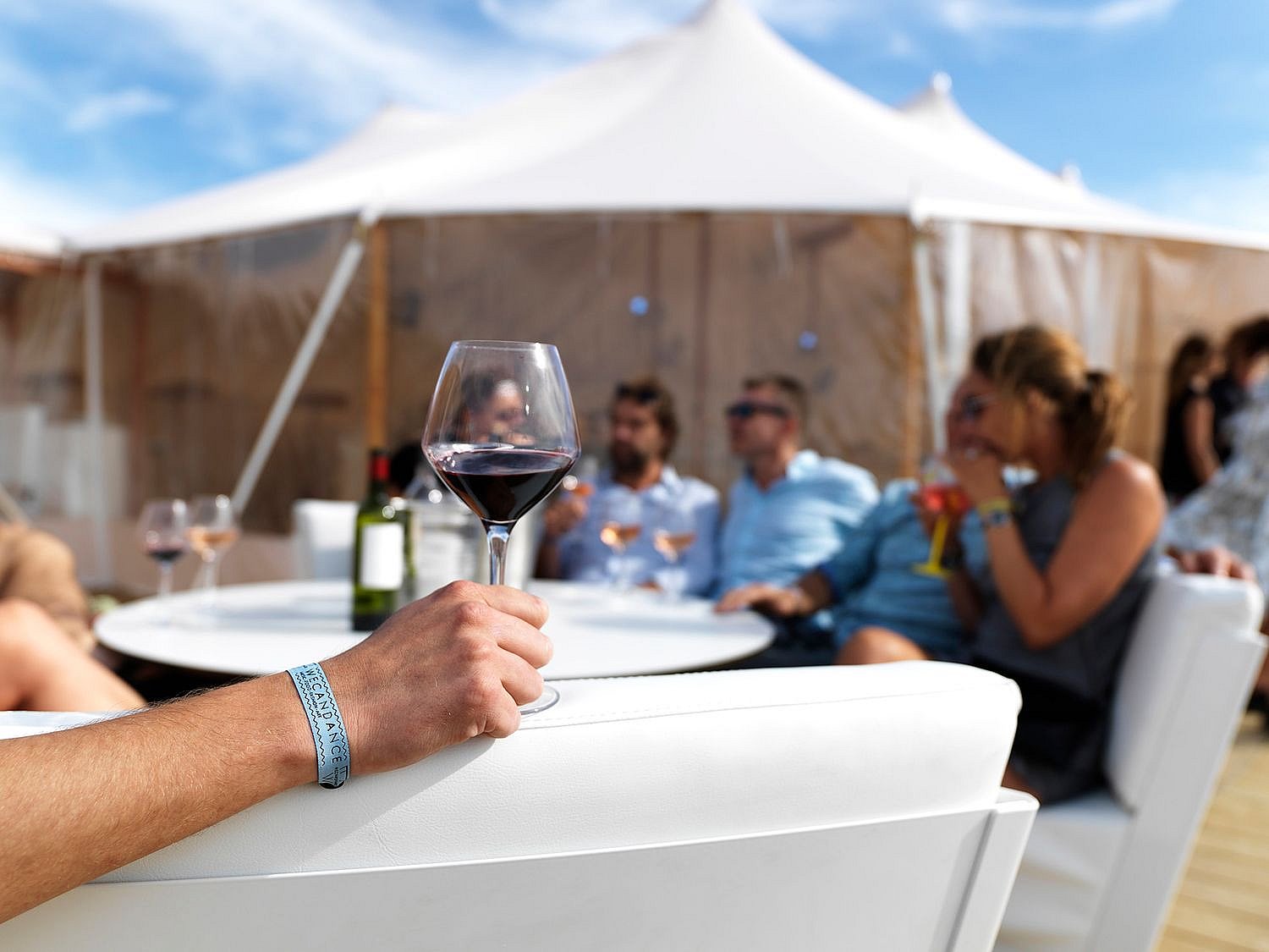   Une main tient un verre de vin rouge tandis que des invités se détendent autour de tables basses blanches, à l'extérieur d'une grande tente, sous un ciel bleu.