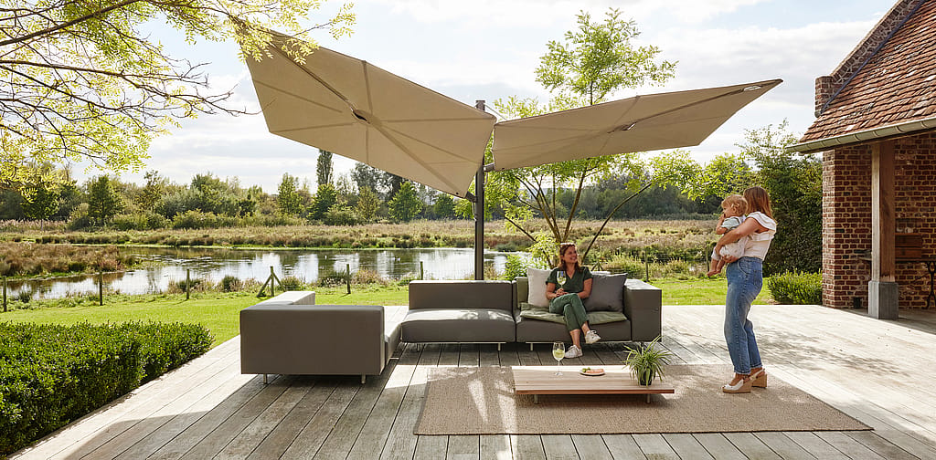 Ein modulares Walrus-Gartensofa steht auf einer Holzterrasse unter großen Sonnenschirmen an einem Fluss. Zwei Frauen und ein Kind genießen die Sitzgelegenheit.