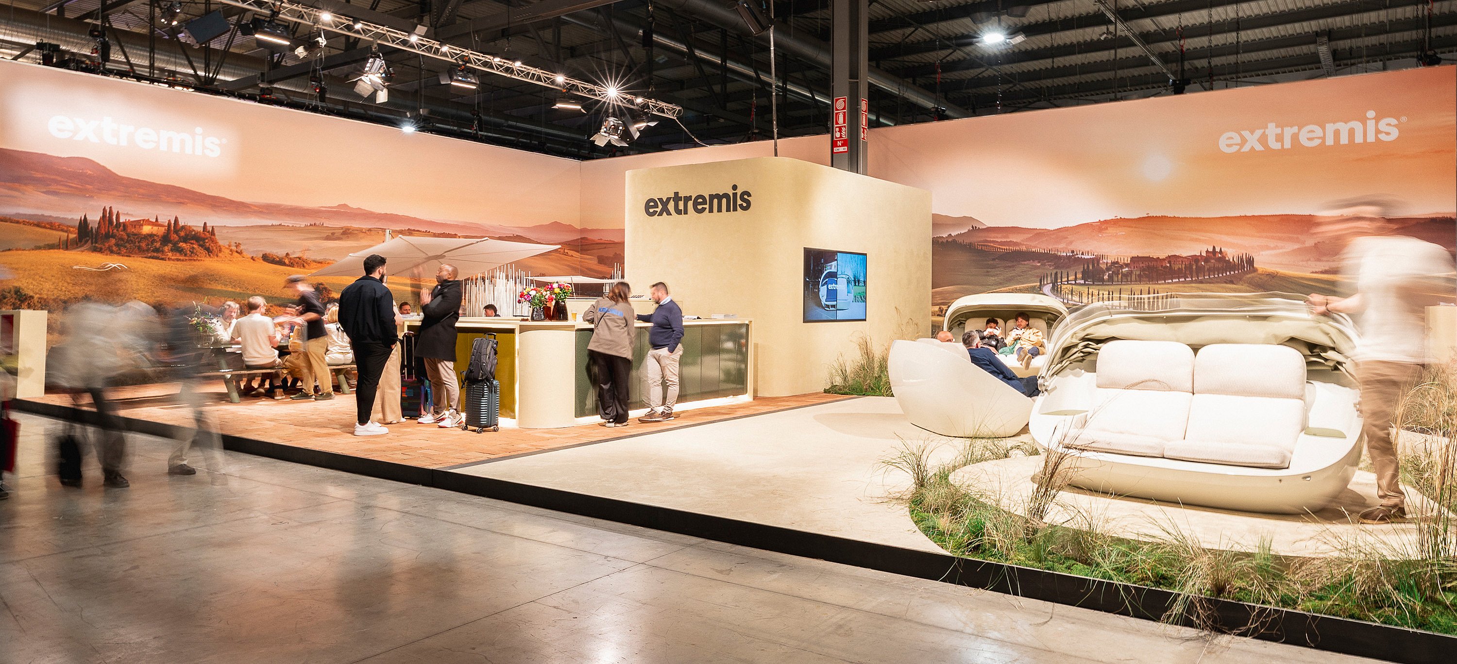 Breed uitzicht op de Extremis-stand in Salone del Mobile, met een panoramische Toscaanse heuvelachtergrond, een receptieteller, bezoekers en Cabrio-banken in een met gras begroeide omgeving.