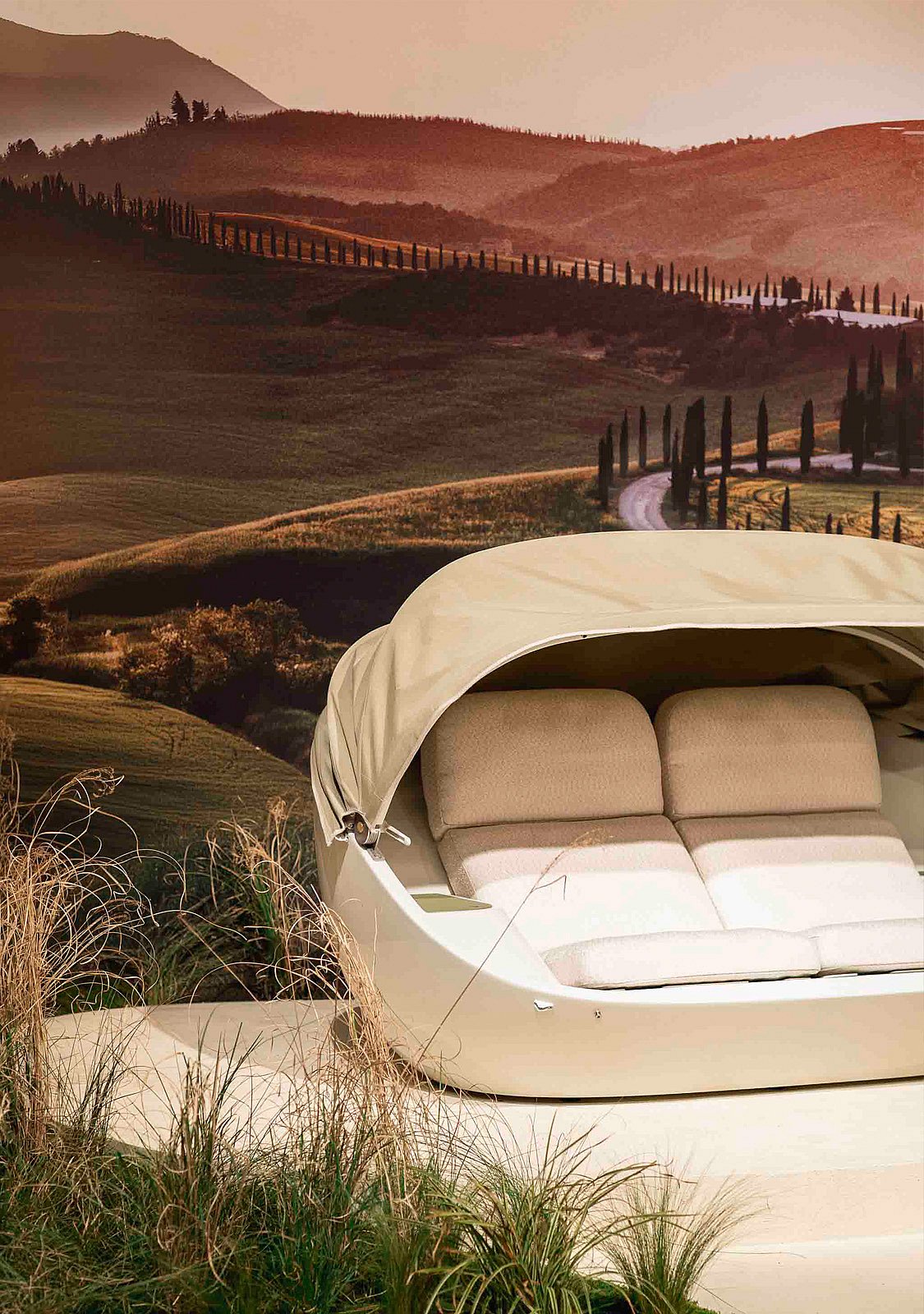 Roomkleurige Cabrio-buitenbank met zachte bovenkant omhoog, geplaatst voor een warme Toscaanse landschapsachtergrond en siergrassen.