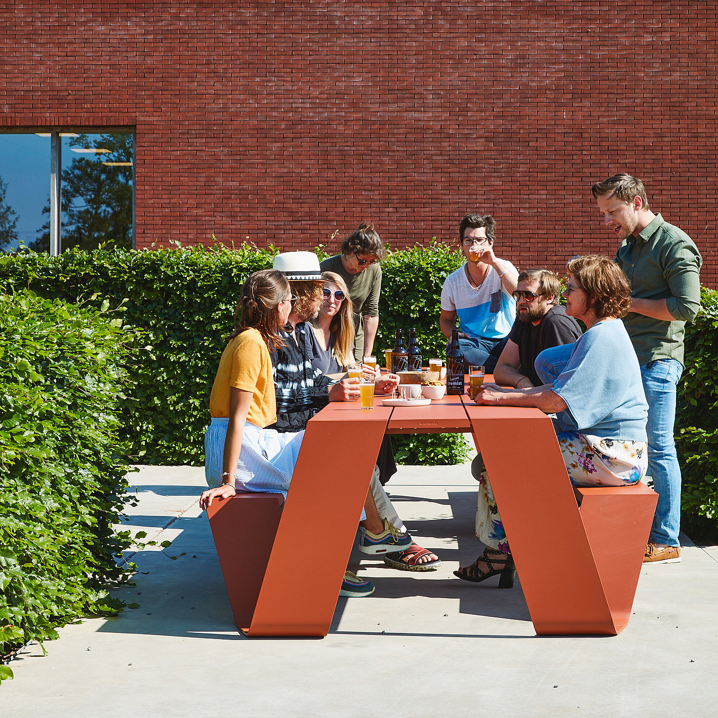 A group of friends sat around a rust-colored picnic table in the garden, drank Tremist beer and chatted in the bright sun next to a hedge and a red brick wall.