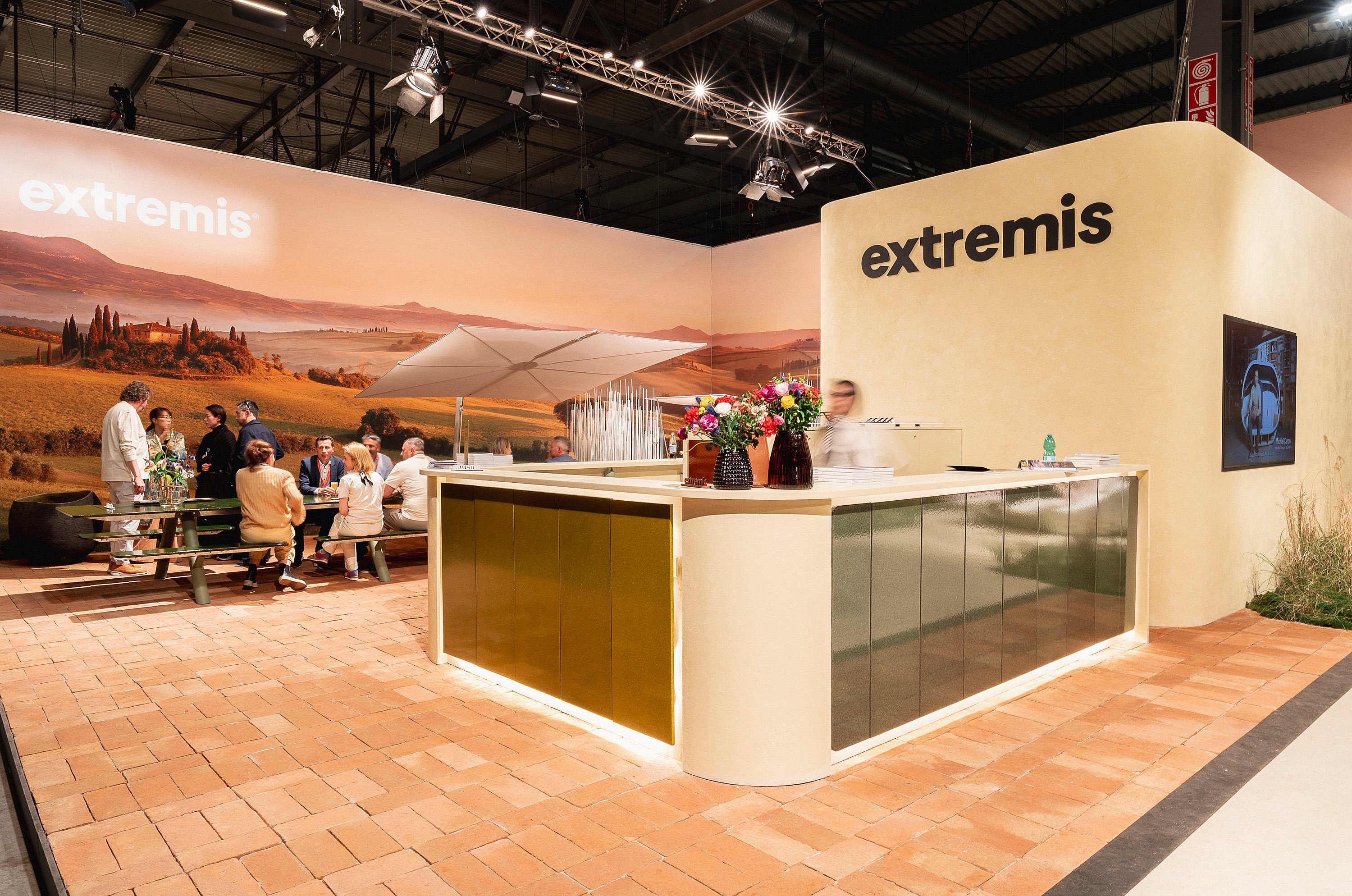 Receptiebalie bij de Extremis-stand met bloemstukken op de voorgrond en bezoekers zitten rond een tafel op de achtergrond.