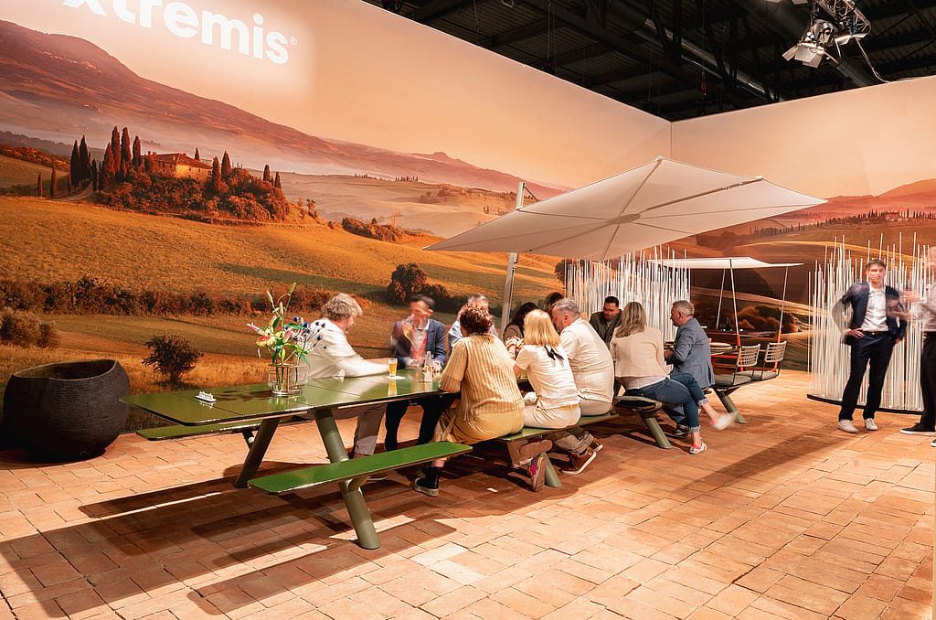 Bezoekers verzamelden zich rond een lange groene Panigiri Lava-tafel met geïntegreerde banken onder een grote witte parasol, tegen een muurschildering van Toscaanse heuvels.