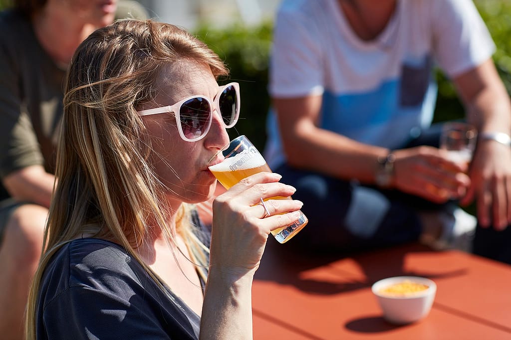 Une femme portant des lunettes de soleil blanches sirote une bière à une table en terrasse ; d'autres personnes sont floues en arrière-plan.