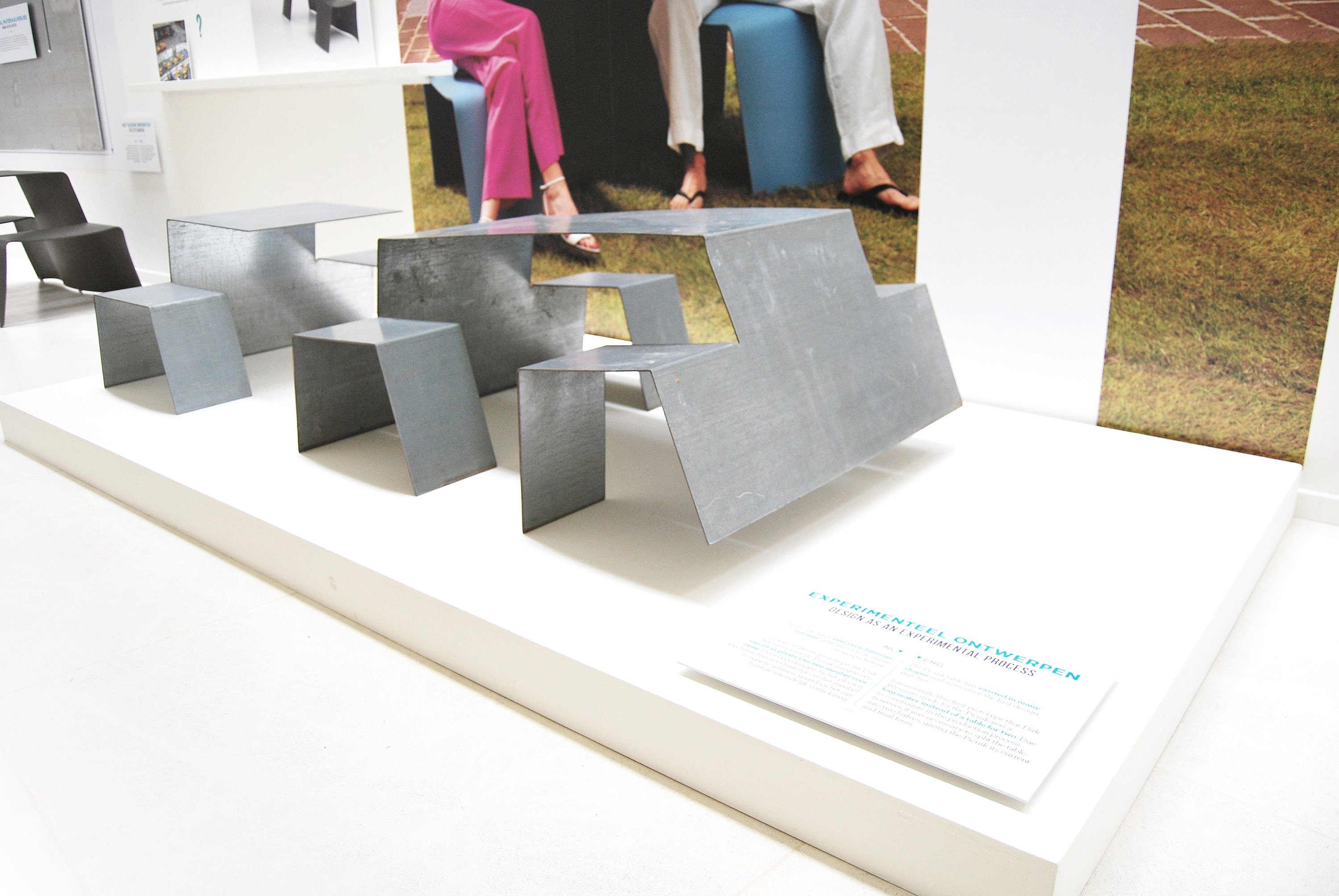 Prototype display of Picnik made from folded sheet metal, shown with small matching stool forms on a white pedestal.
