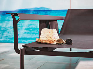 Nahaufnahme des Sol+Luna Outdoor-Sofas oder Sonnenbetts in dunklem Finish neben hellblauem Wasser, mit Strohhut und Sonnenbrille auf dem Sitz.