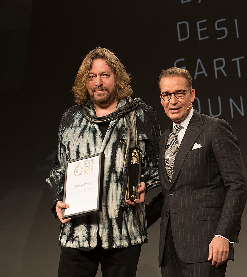 Dirk Wynants se tient sur scène, tenant le trophée et le certificat du German Design Award à côté d’un homme en costume sombre lors de la cérémonie de remise des prix à Francfort.