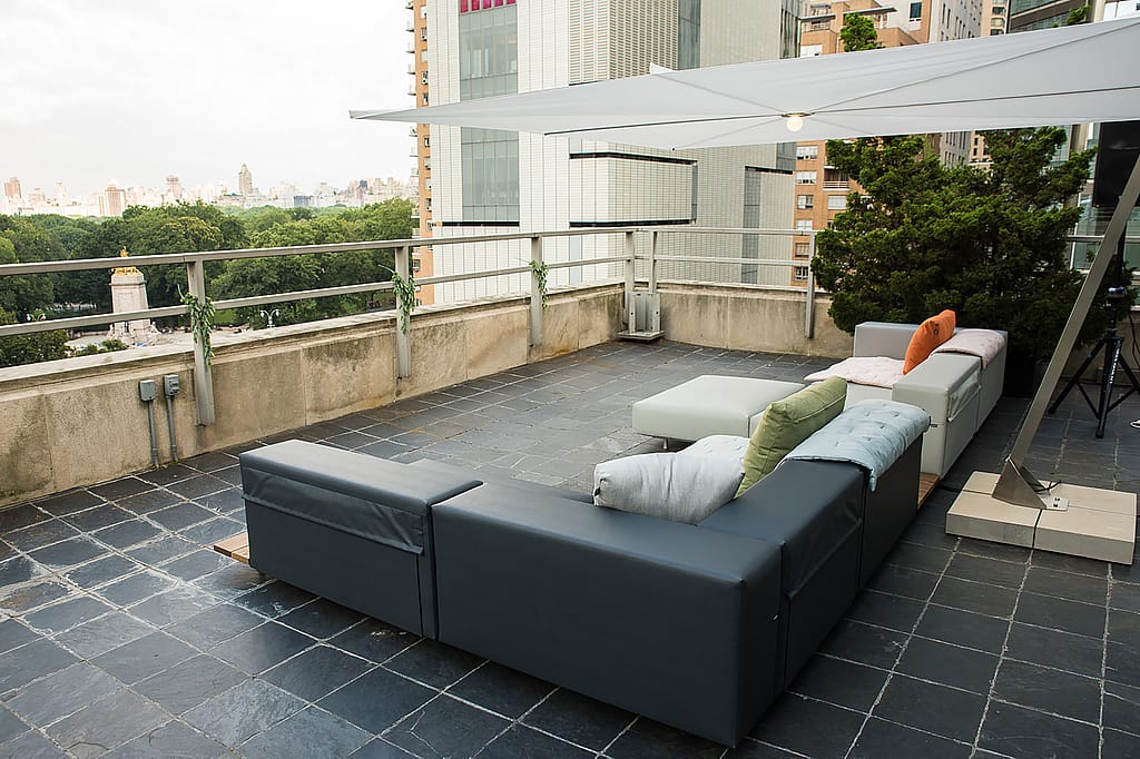 Grand salon extérieur modulable avec des coussins pâles sous une voile d’ombrage blanche étirée sur le toit-terrasse.