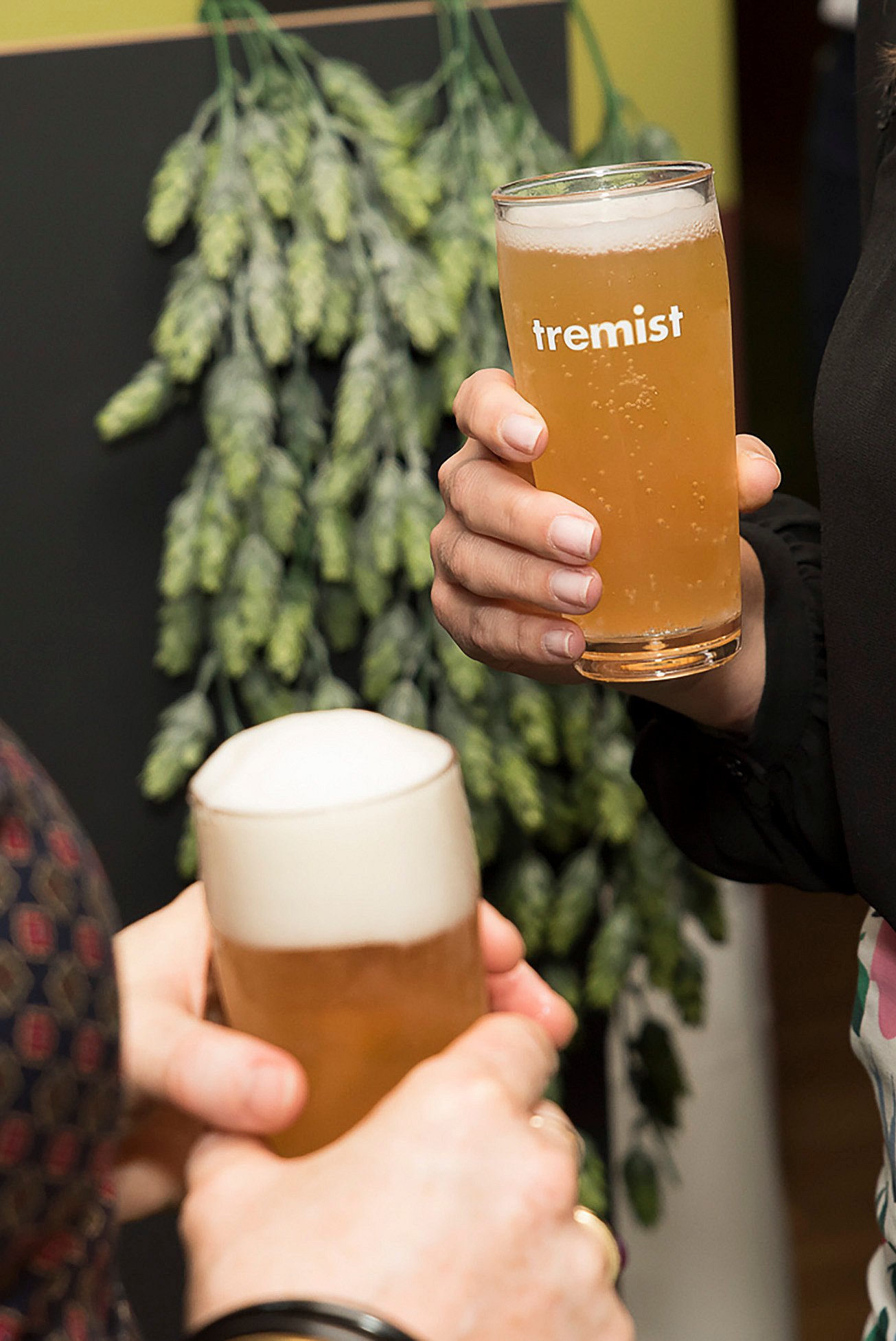 Gros plan d’une main tenant un verre de bière pâle marqué « tremist », avec une verdure floue en arrière-plan.