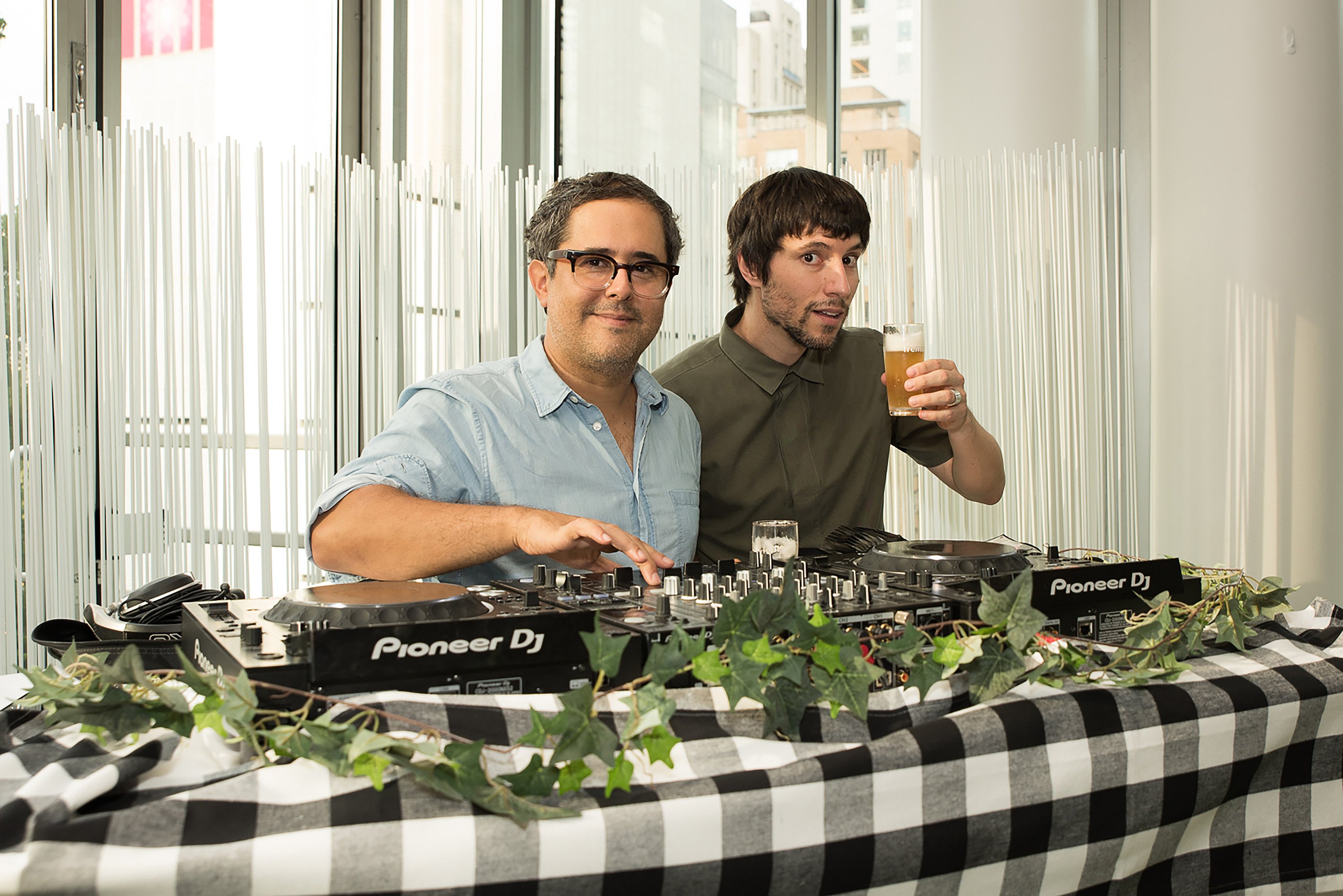 Deux DJs derrière une cabine de DJ à carreaux décorée de lierre, l’un regardant vers la caméra tandis que l’autre tient une boisson.