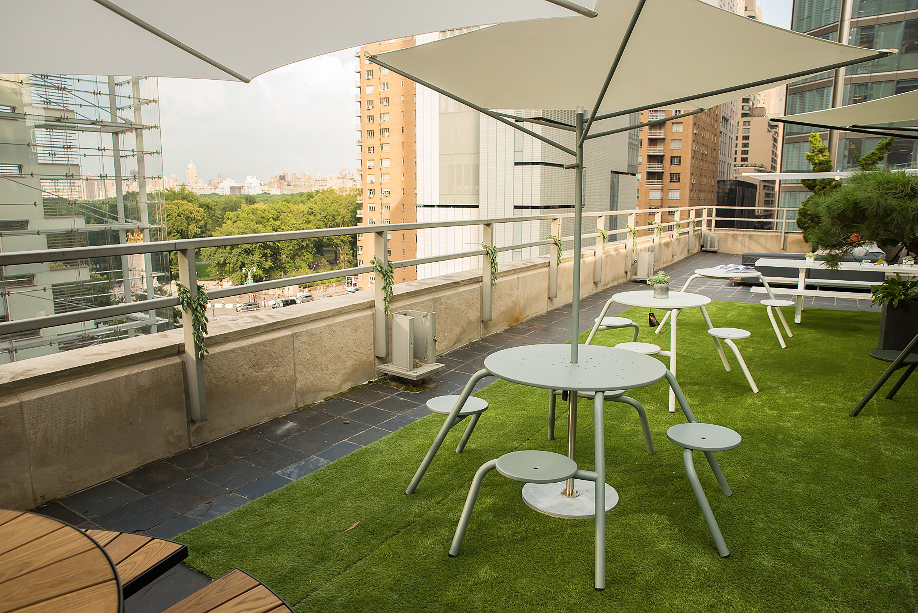 Tables et tabourets minimalistes de café blanc disposés sur du gazon artificiel sous de grands parasols sur la terrasse du toit, avec les bâtiments de la ville et Central Park au-delà.