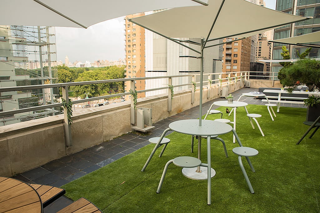 Tables et tabourets minimalistes de café blanc disposés sur du gazon artificiel sous de grands parasols sur la terrasse du toit, avec les bâtiments de la ville et Central Park au-delà.