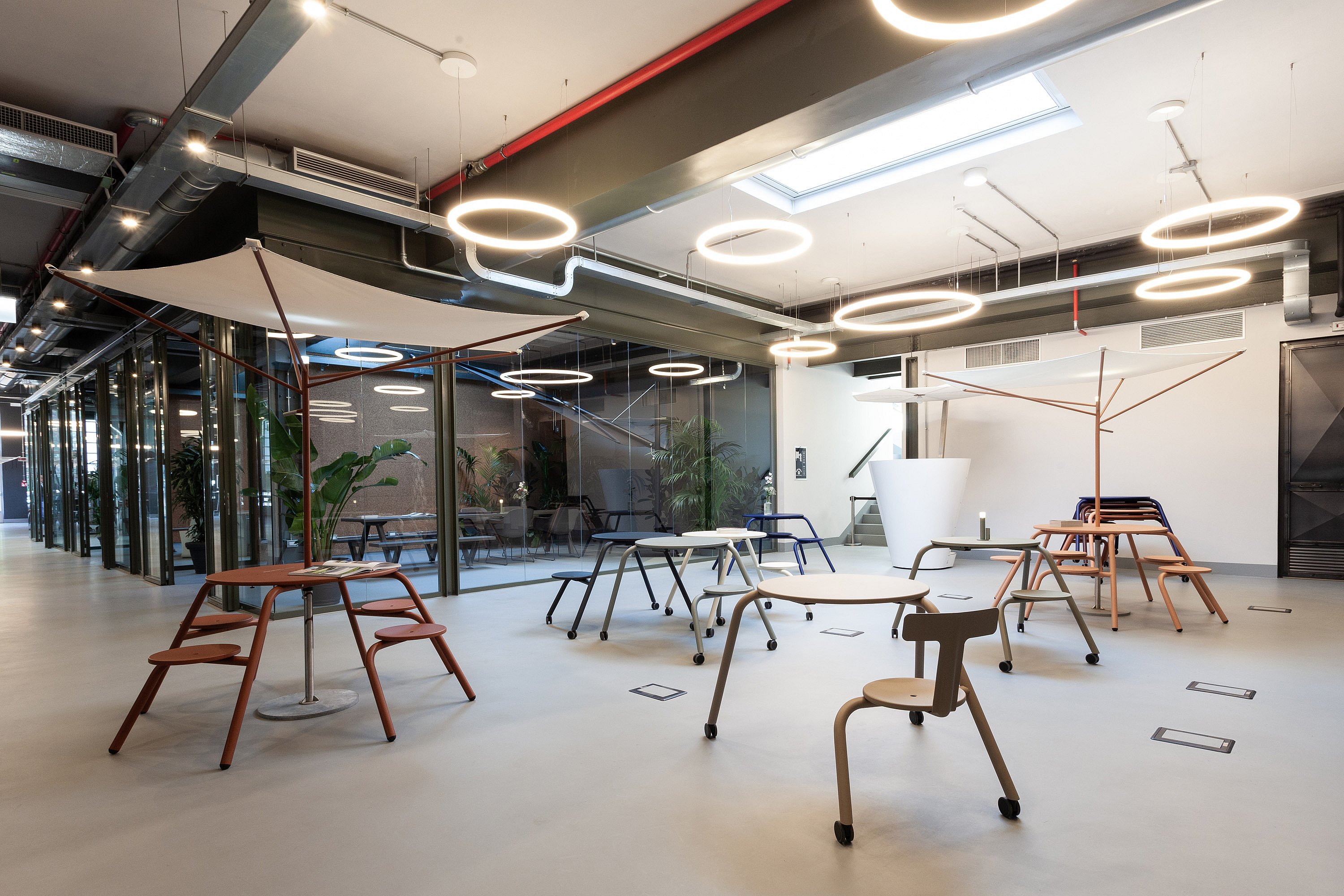 Vue d'ensemble du showroom avec des tables, des tabourets, des bancs et des parasols colorés disposés dans un espace ouvert sous des anneaux lumineux.
