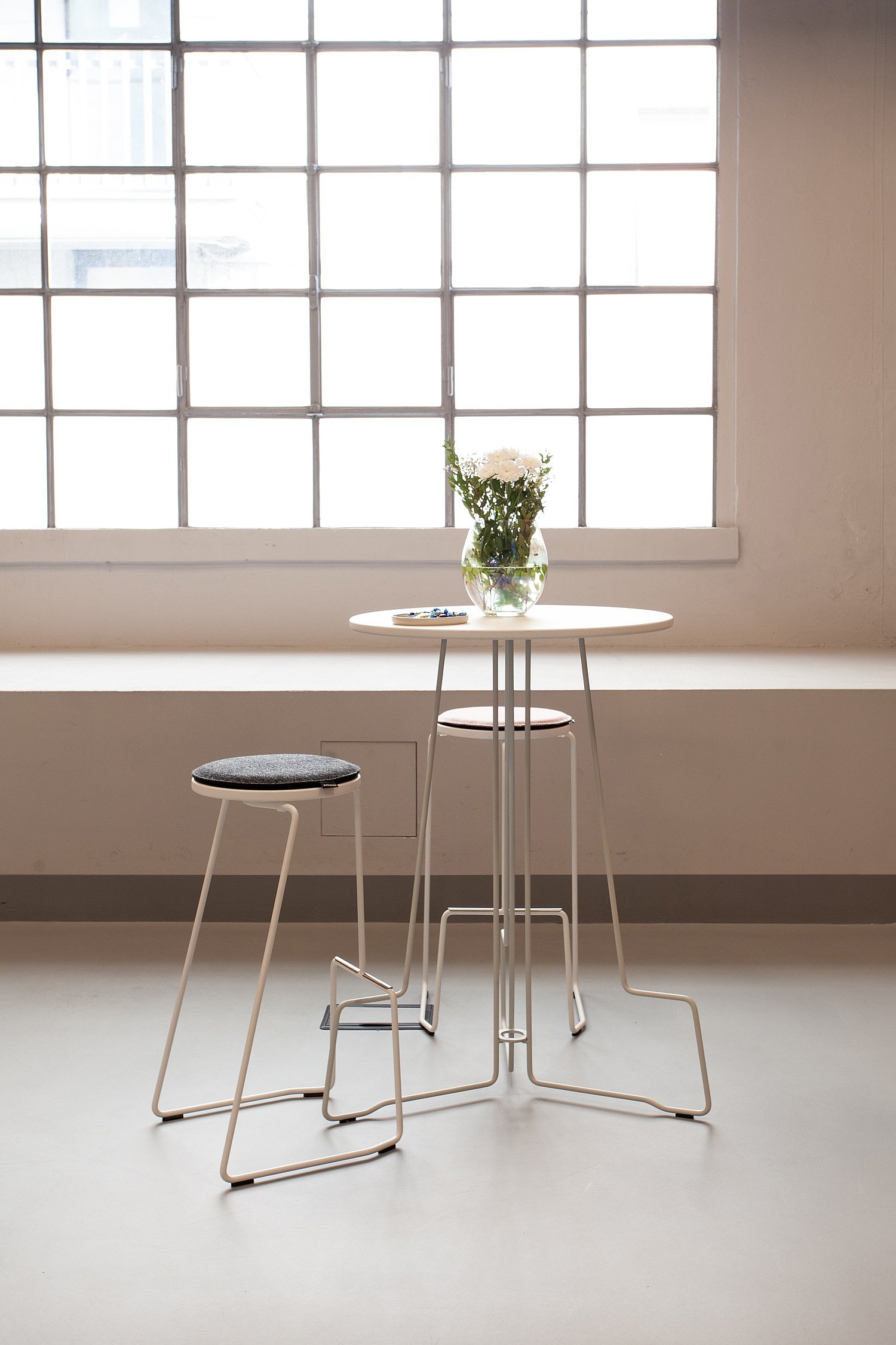Une table ronde blanche minimaliste, à hauteur standard, avec deux tabourets près d'une grande fenêtre, surmontée d'un vase en verre contenant des fleurs blanches.