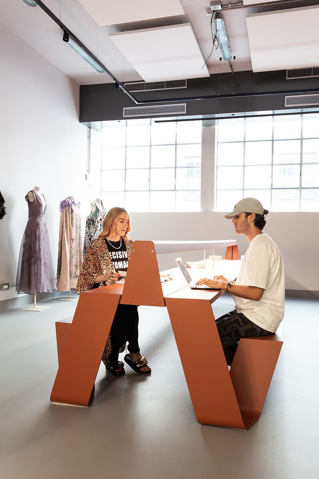 Deux personnes travaillent face à face à une table couleur rouille avec des panneaux de séparation inclinés, dans une pièce lumineuse avec des mannequins et des robes en arrière-plan.
