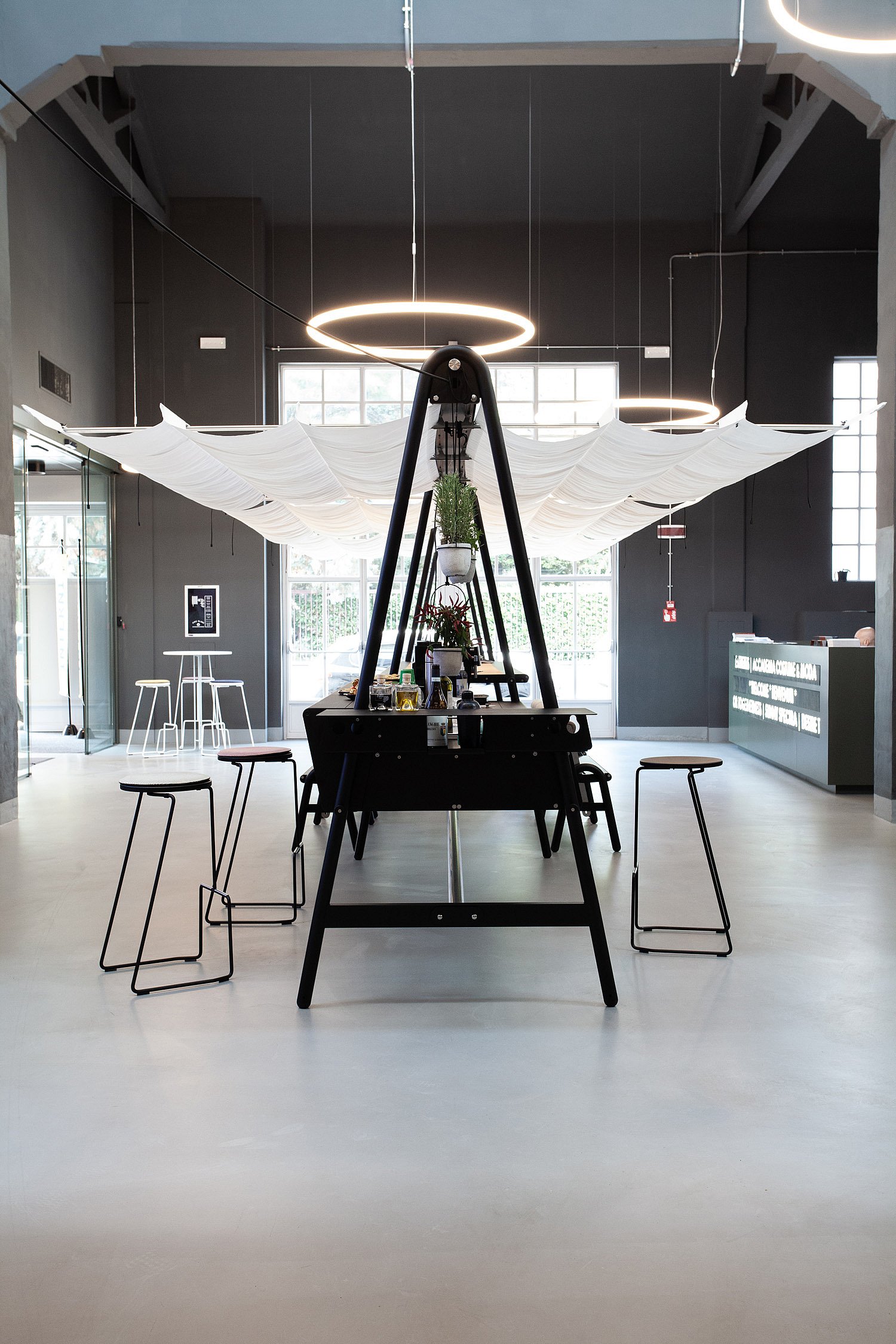 Vue de face d'un long ensemble table et banc AMAi noirs, installé dans un hall moderne, avec des panneaux d'ombrage blancs drapés au plafond, des luminaires annulaires, des tabourets et une plante suspendue au centre.