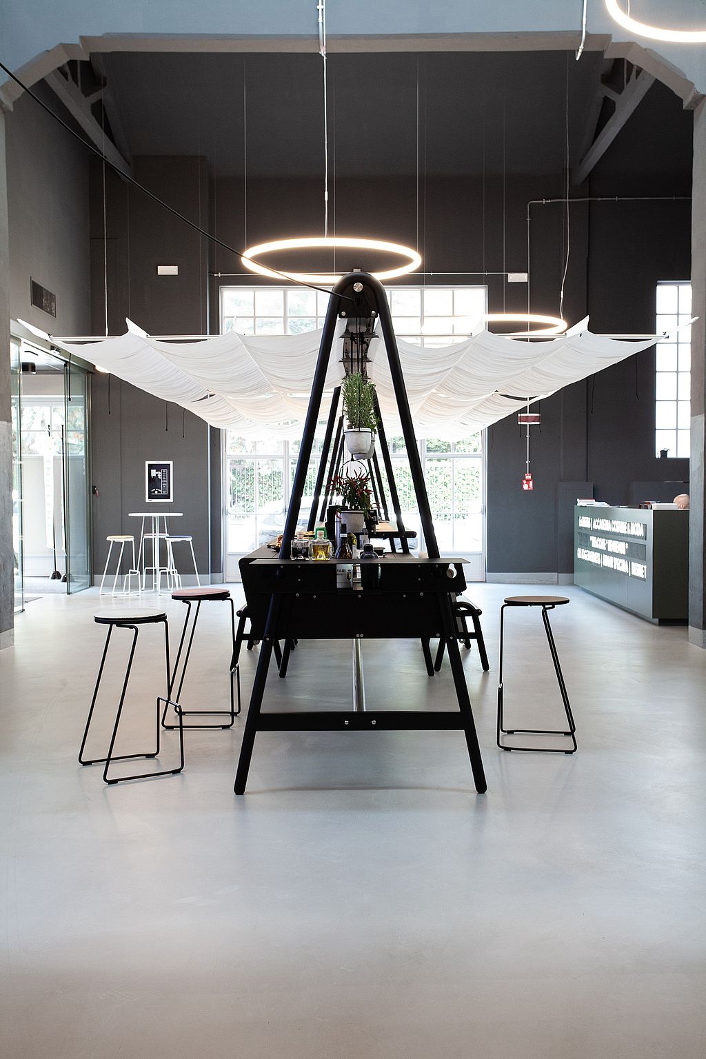 Vue de face d'un long ensemble table et banc AMAi noirs, installé dans un hall moderne, avec des panneaux d'ombrage blancs drapés au plafond, des luminaires annulaires, des tabourets et une plante suspendue au centre.