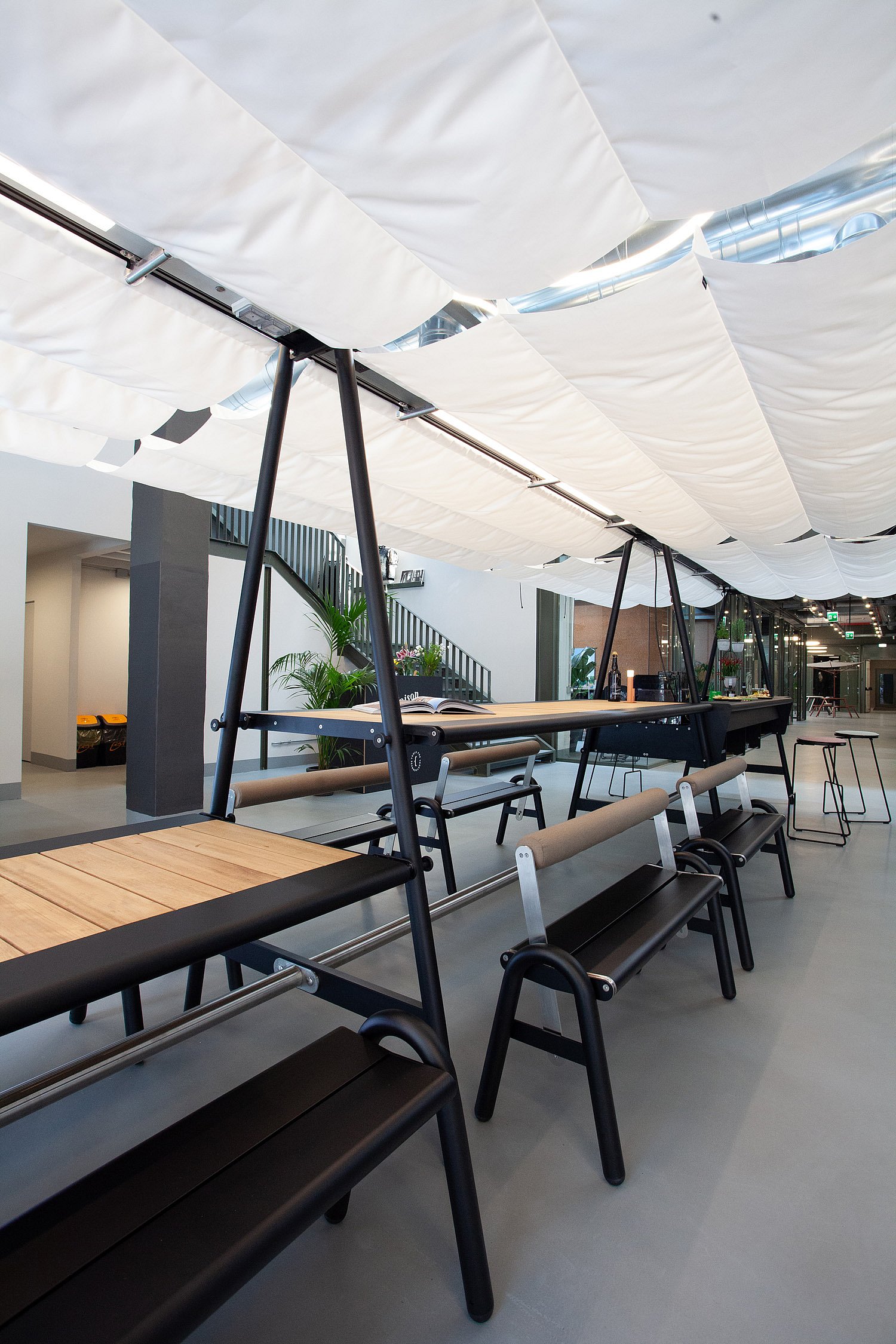 Vue en perspective de plusieurs tables et bancs AMAi noirs alignés à l'intérieur, sous de longs panneaux de tissu blanc suspendus.