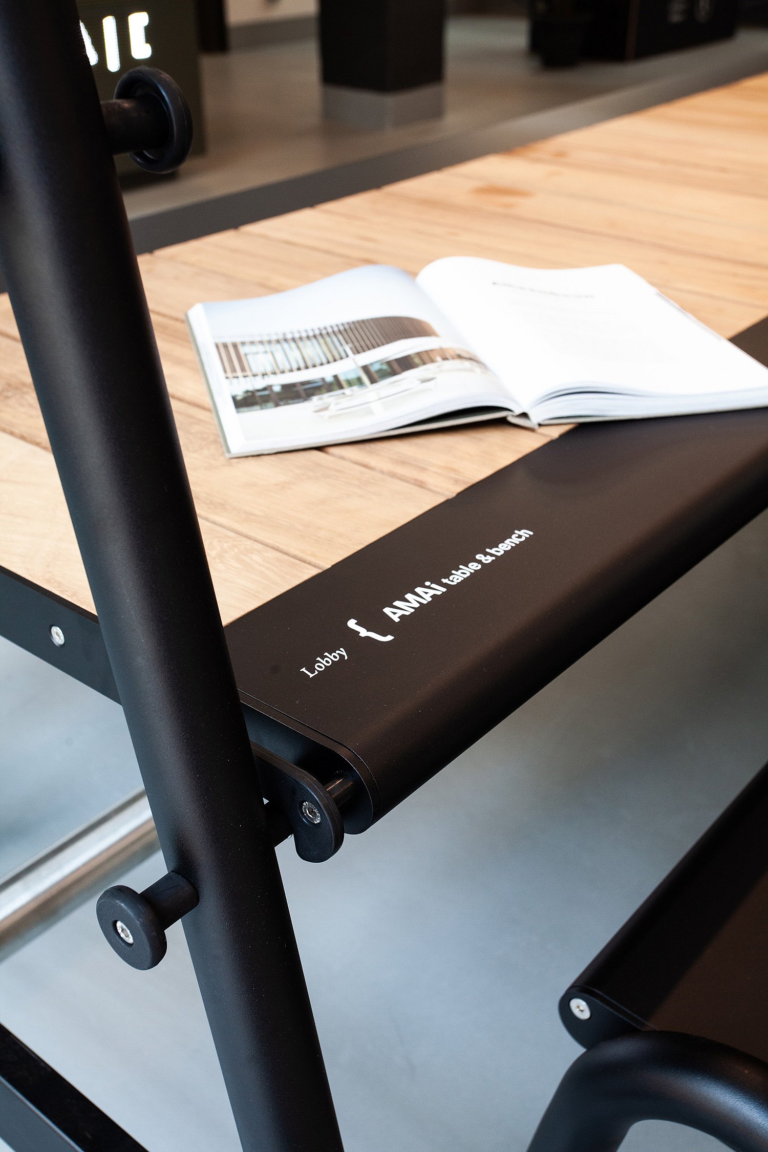 Gros plan sur un plateau de table en bois avec un livre ouvert posé sur une structure métallique noire portant l'inscription « Table et banc AMAi du hall ».