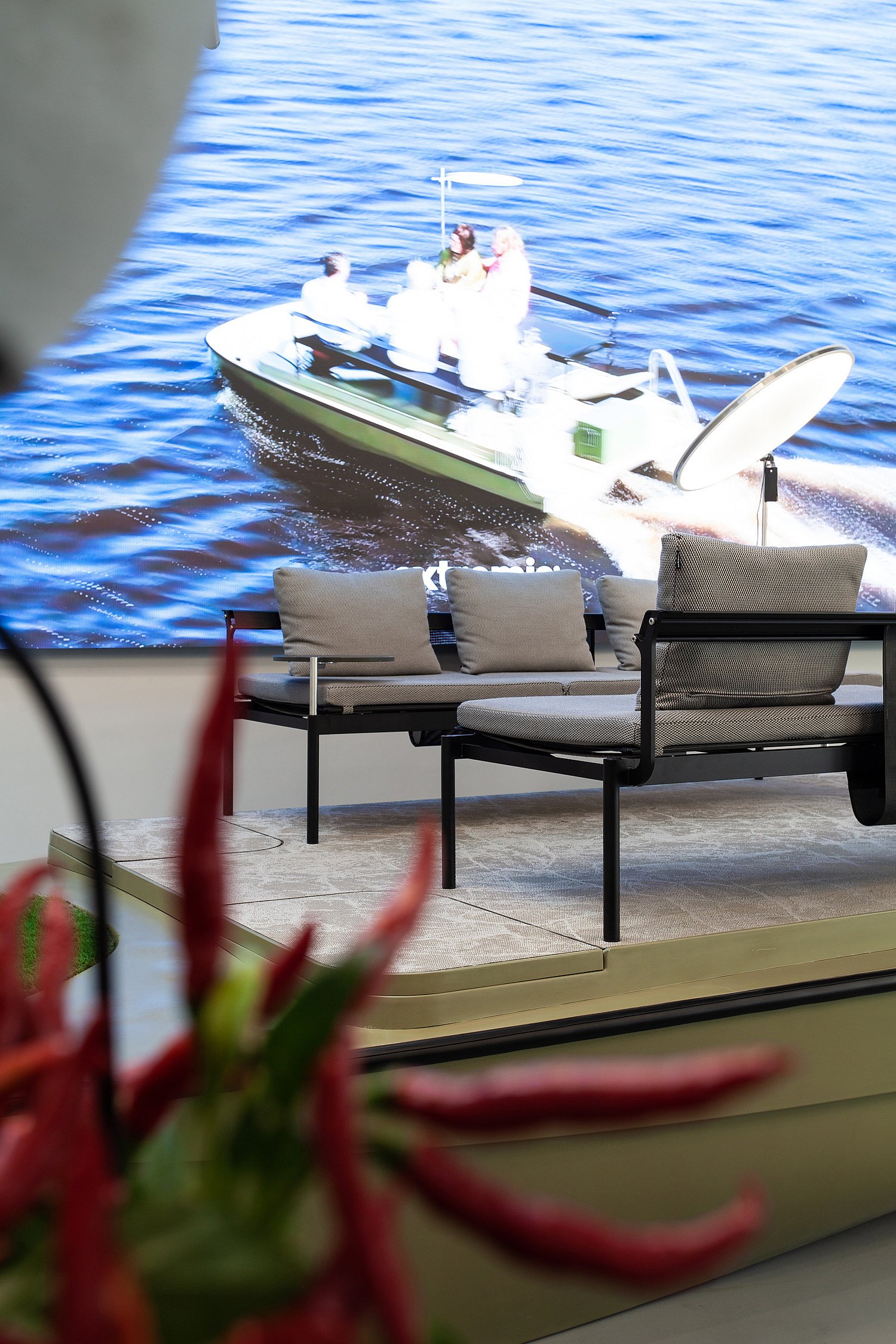   Gros plan sur un canapé d'extérieur à cadre noir et une table ronde disposés sur le bateau, avec un grand écran représentant un paysage aquatique en arrière-plan et des fleurs rouges floues au premier plan.