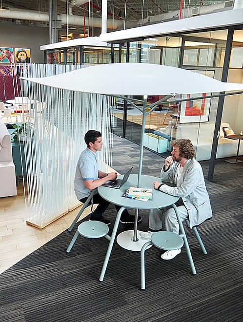 Indoor Acacia-stijl akoestische luifel met een brede bleke stoffen bovenkant boven een ronde tafel en krukken, in een open kantoorlounge met zachte stoelen en planken op de achtergrond.