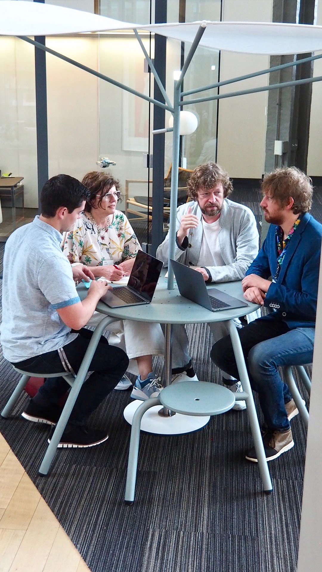 Vier collega's verzamelen zich rond een ronde tafel met open laptops onder het vertakte Acacia-frame en de luifel, met de opstelling als ontmoetingsplek binnenshuis.