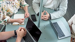 Vier collega's verzamelen zich rond een ronde tafel met open laptops onder het vertakte Acacia-frame en de luifel, met de opstelling als ontmoetingsplek binnenshuis.