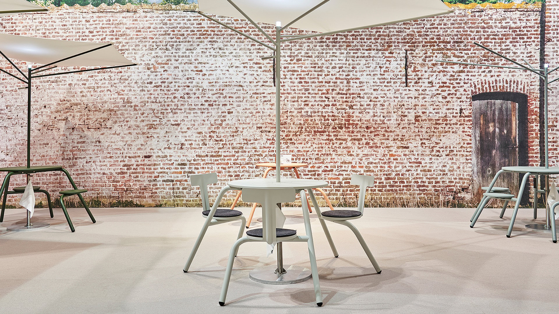 Table ronde blanche et sièges sous le parasol sculptural Acacia, contre un mur de briques dans le stand d’exposition.