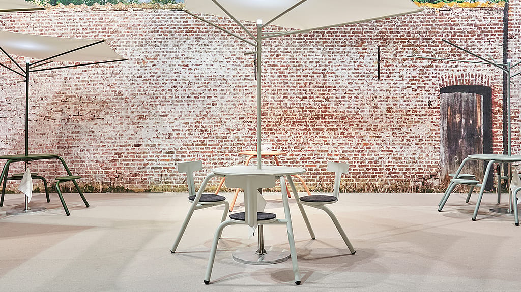 Table ronde blanche et sièges sous le parasol sculptural Acacia, contre un mur de briques dans le stand d’exposition.