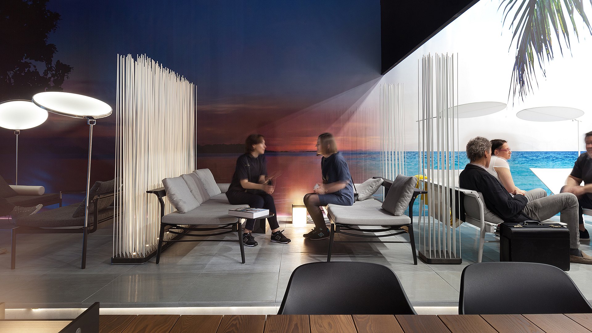 Deux personnes assises sur des canapés gris Sol + Luna sous un décor de coucher de soleil spectaculaire, avec la structure sculpturale du parasol blanc Kosmos entourant en partie l’espace salon.